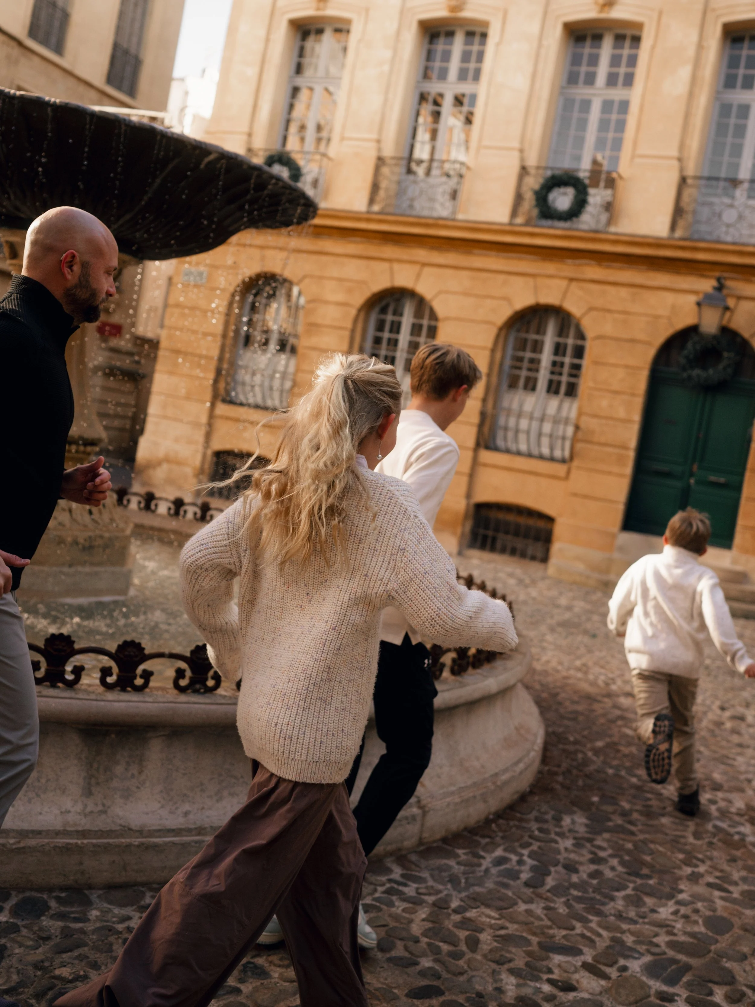 Four people running past a stone fountain on a cobblestone street in front of a European-style building with large windows and a green door.