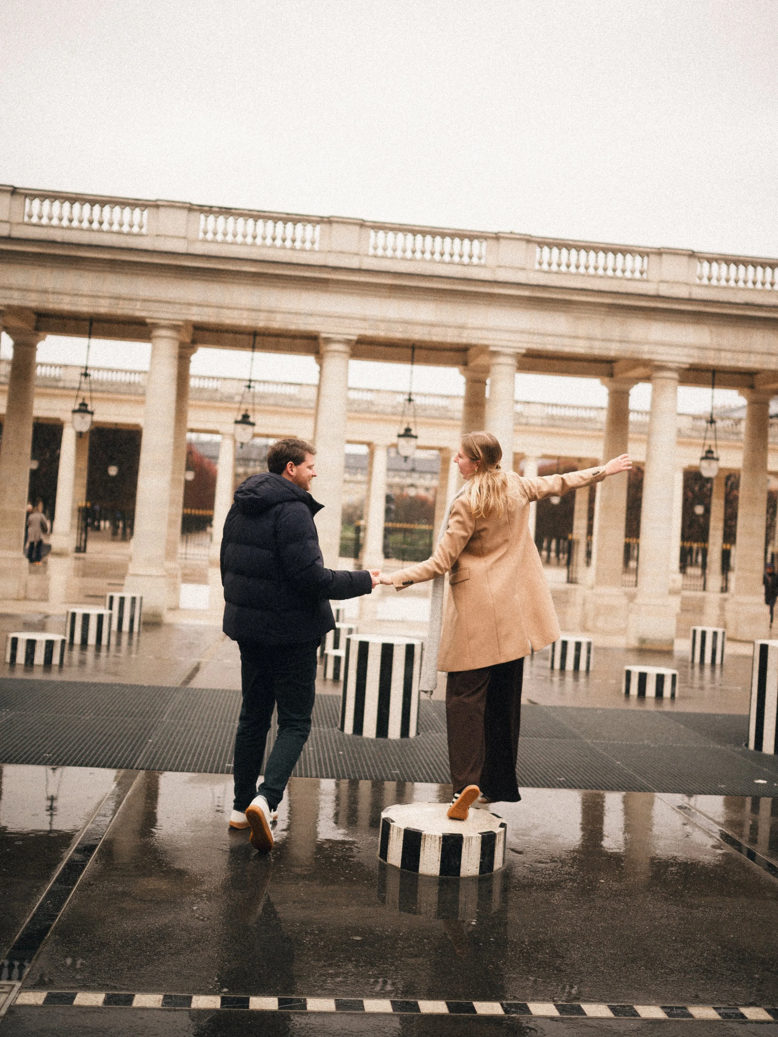 A man and woman holding hands, dancing on striped black and white circular platforms outdoors in an architectural setting with large columns and hanging lamps.