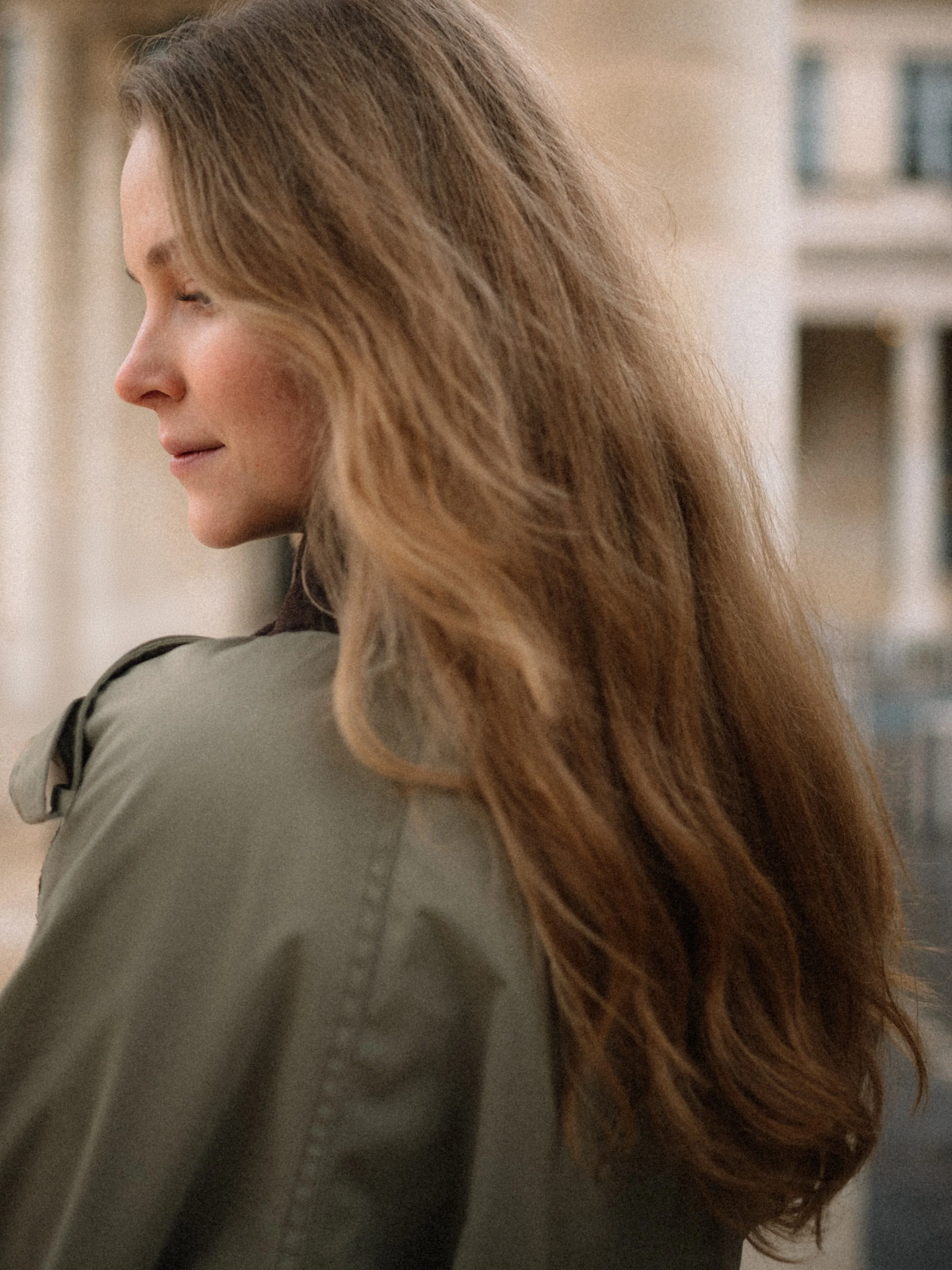 Close-up of a woman with long, wavy brown hair, wearing a green jacket, with an urban background of buildings.