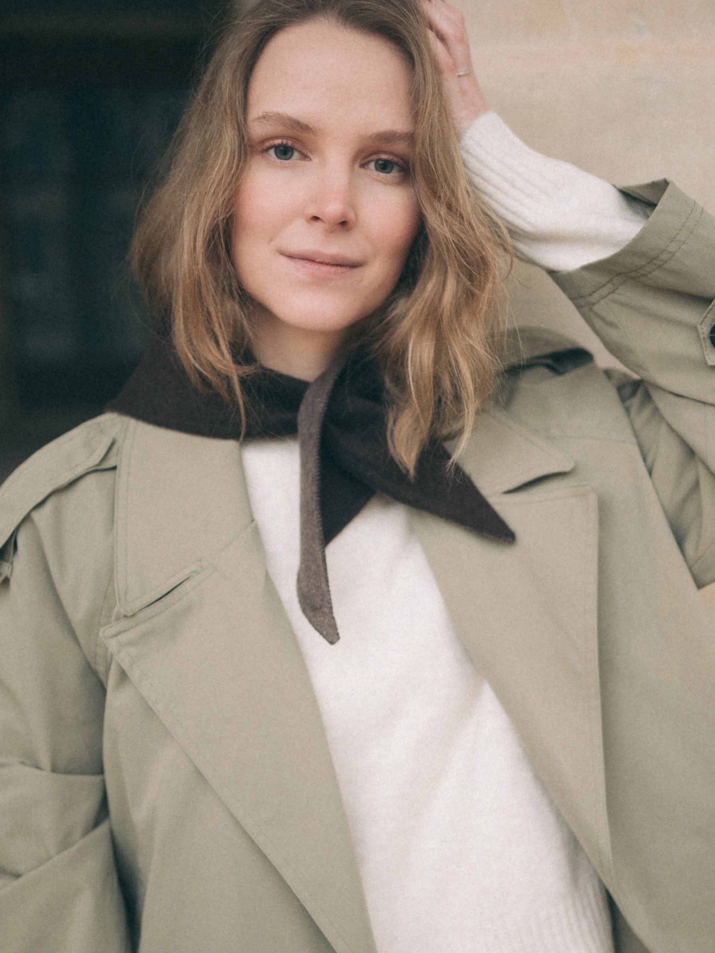 A young woman with wavy light brown hair, light skin, and blue eyes, wearing a beige trench coat over a white sweater and a black scarf, smiling and posing with her hand behind her head.