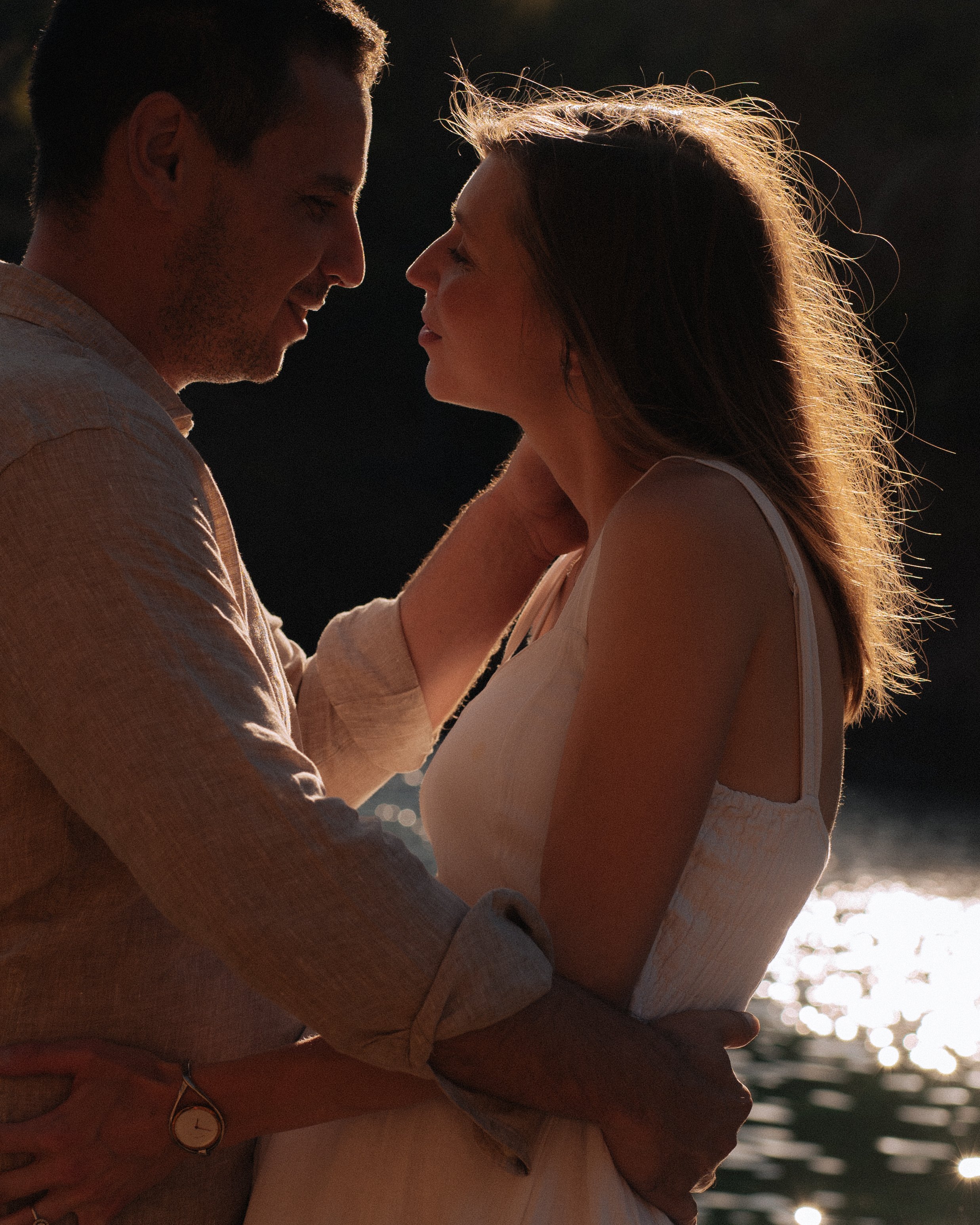 A couple stands close together by water at sunset, gazing into each other's eyes with the sun behind them creating a silhouette effect.