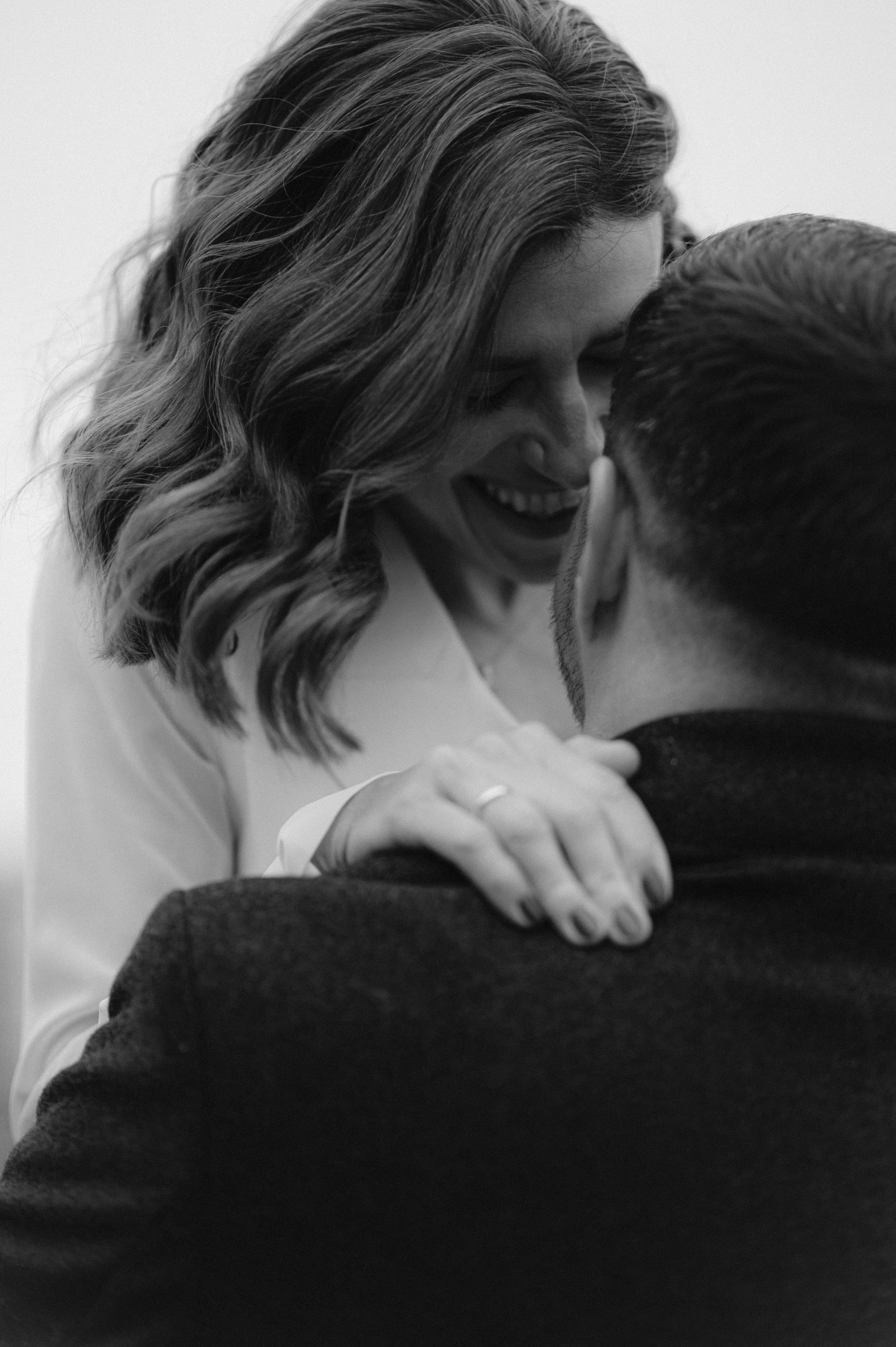 Black and white photo of a woman with wavy hair smiling and embracing a man, holding his shoulder while leaning in close.