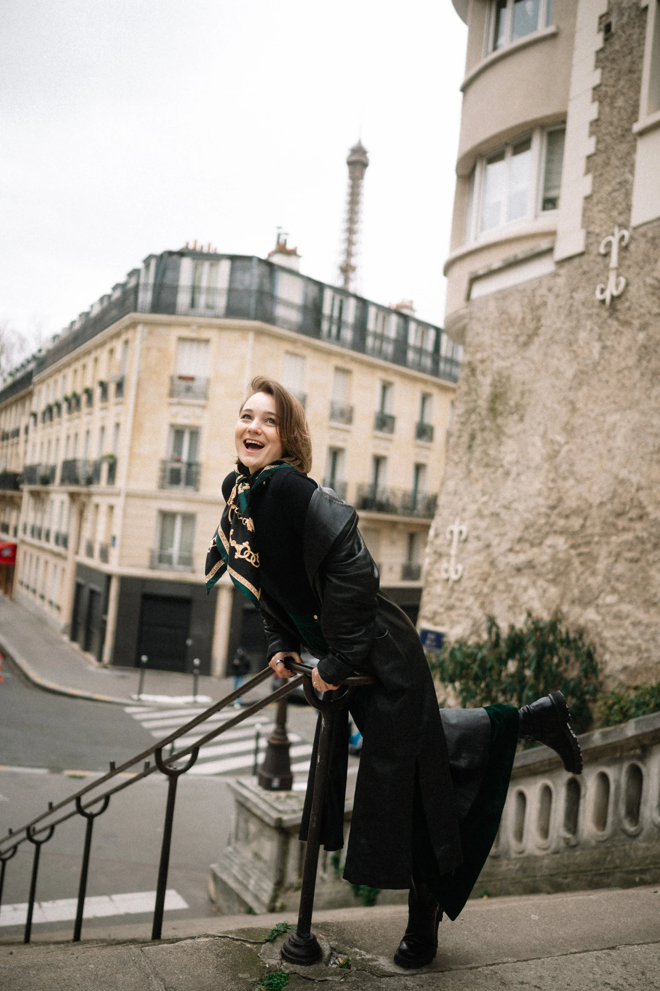 Young woman with short brown hair laughing while balancing on a stair railing in an urban neighborhood with apartment buildings and a tower in the background.