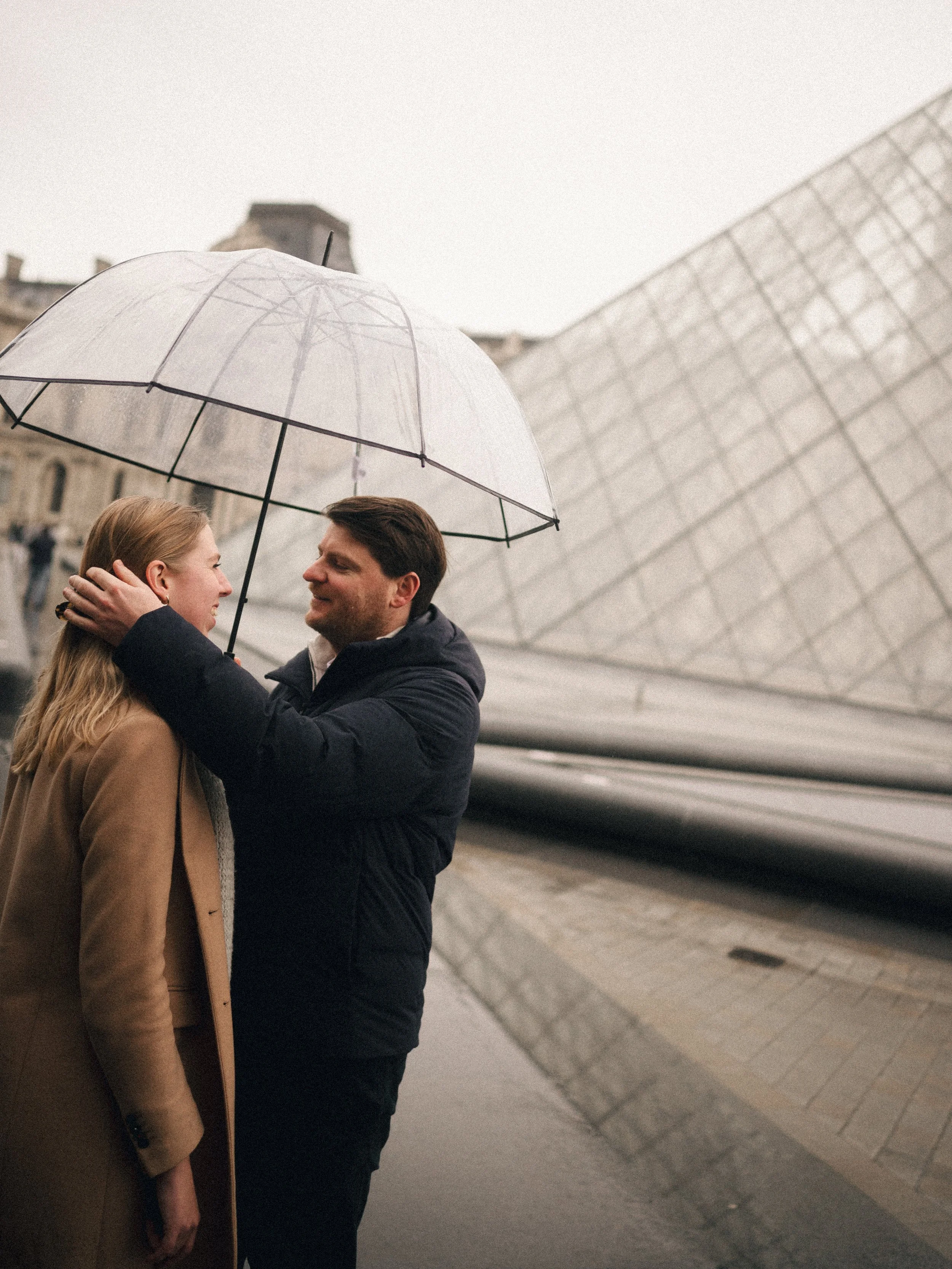 A couple standing close together under a transparent umbrella in front of a modern building on a rainy day. The man is holding the umbrella and touching the woman's face gently.