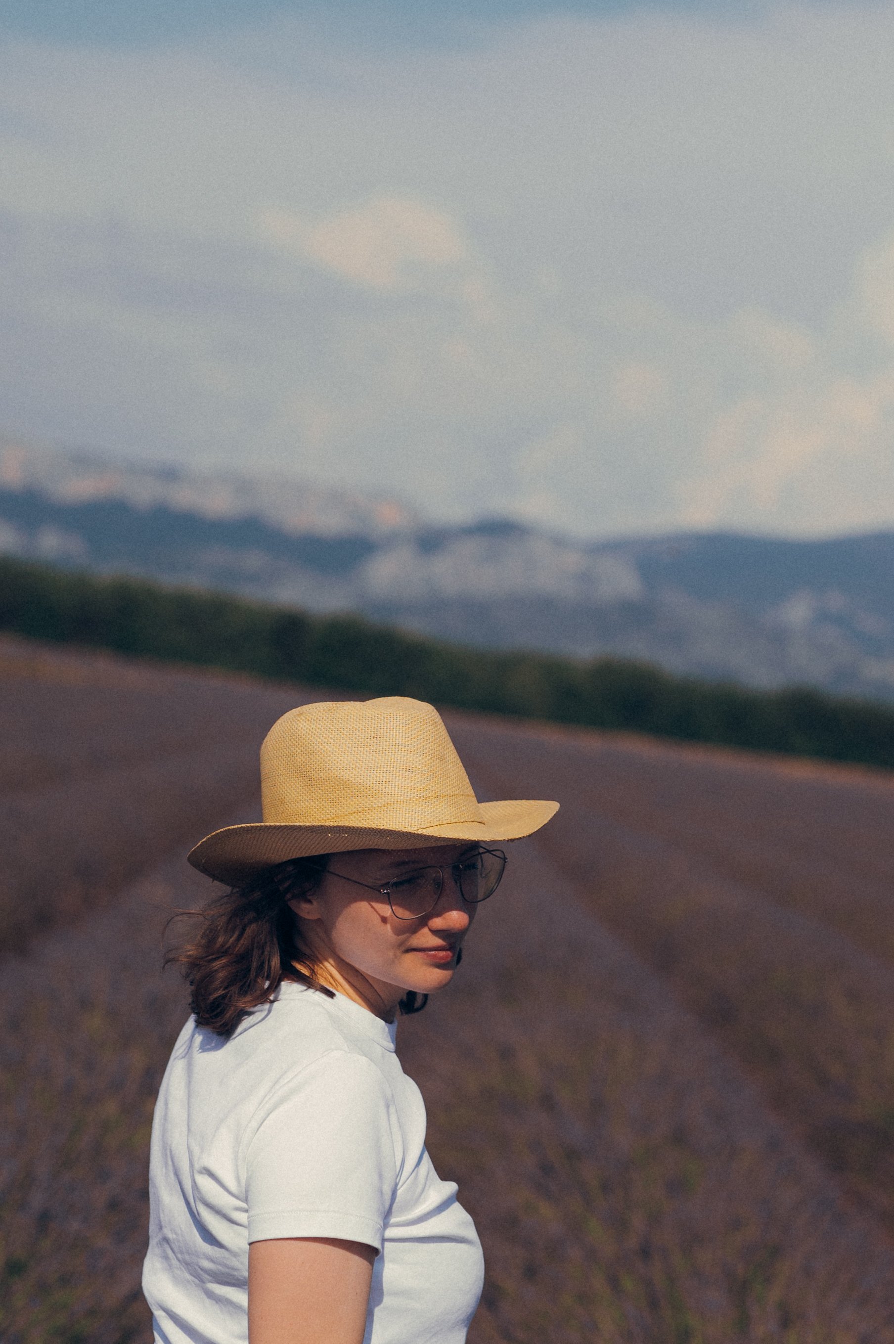 Woman wearing a straw hat and glasses standing in a field with mountains in the background.