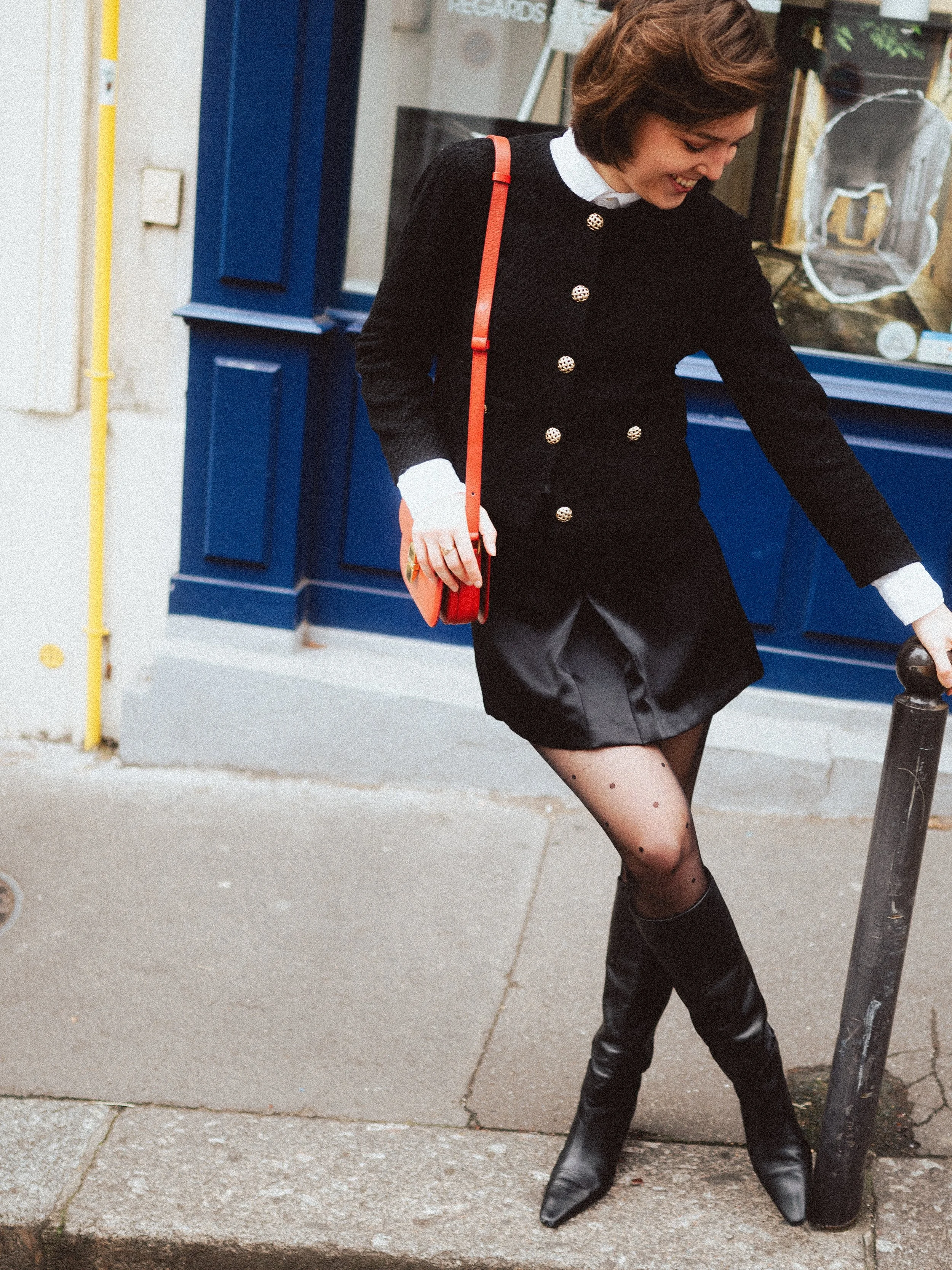 A woman smiling while standing on a sidewalk, wearing a black coat with gold buttons, a short skirt, patterned tights, and tall black boots. She has a small red purse with a shoulder strap and is reaching down to hold a street bollard beside her.