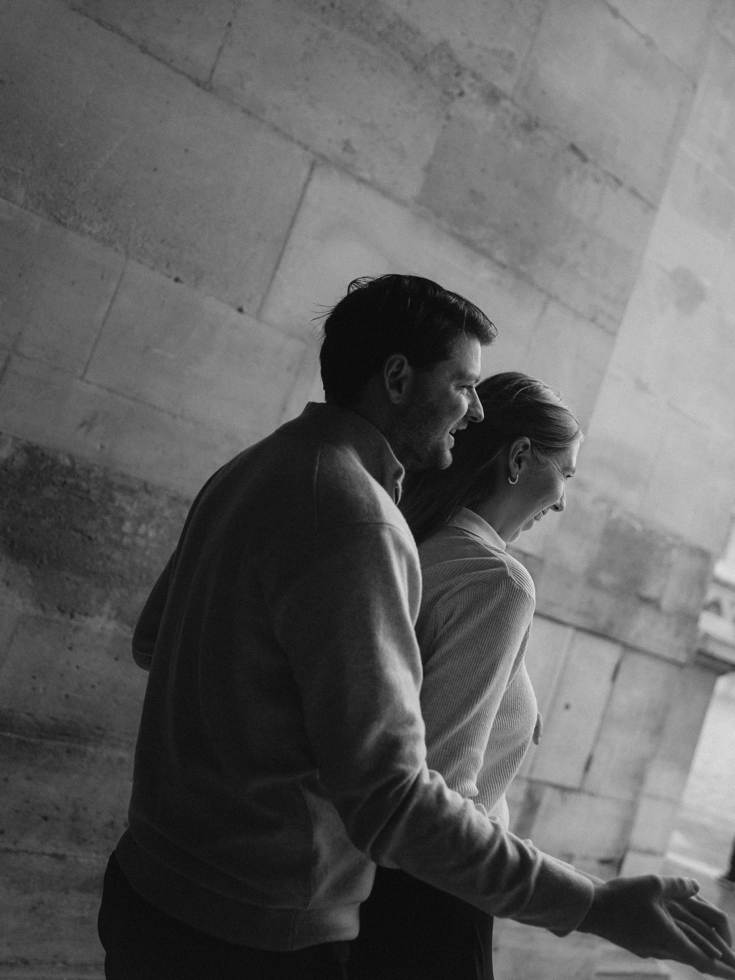 A black and white photo of a smiling man and woman standing side by side against a stone wall, both with their eyes closed, appearing happy and relaxed.