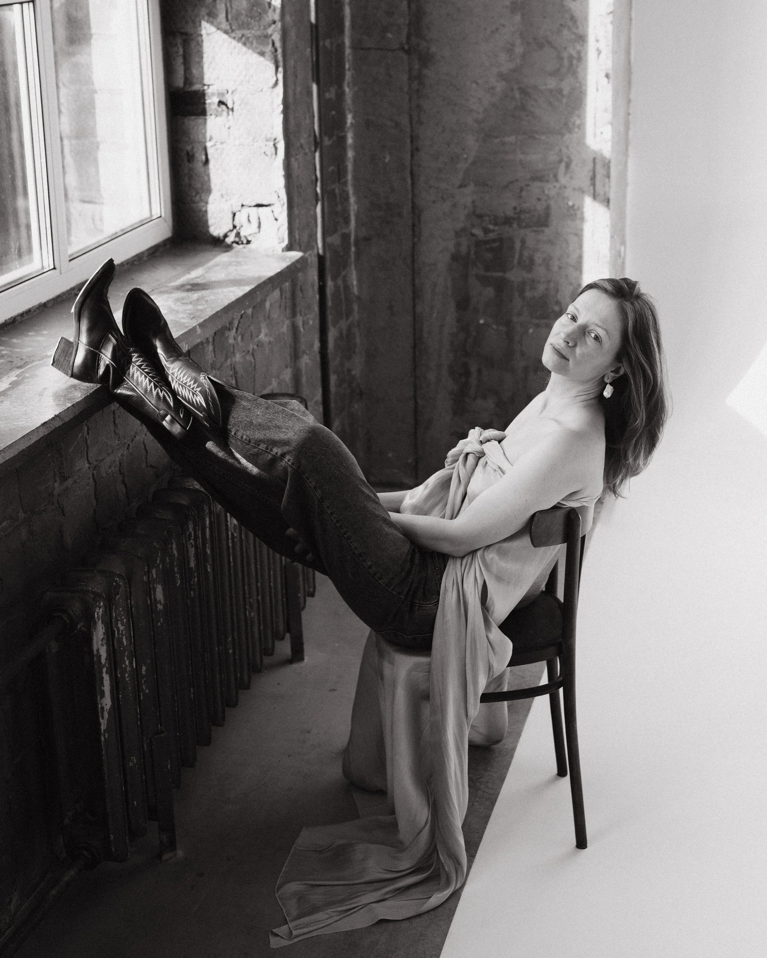 A woman with light skin sitting on a chair with her legs resting on a windowsill, wearing jeans and pointed-toe boots, in an industrial-style room with brick wall and radiator, with a serious expression.