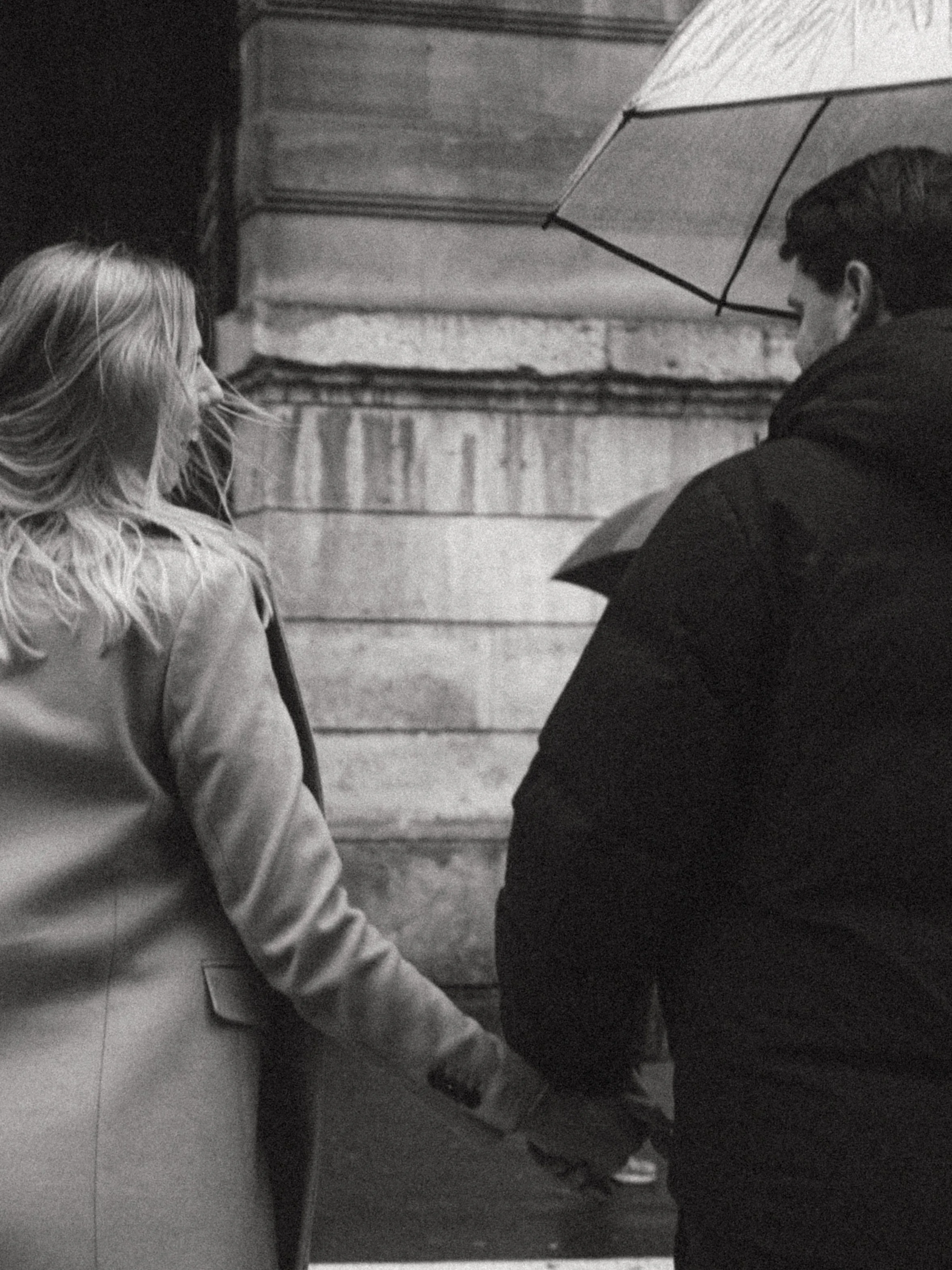 A black and white photo of a couple holding hands and walking together at night, with the woman on the left and the man on the right holding an umbrella, against a stone building background.