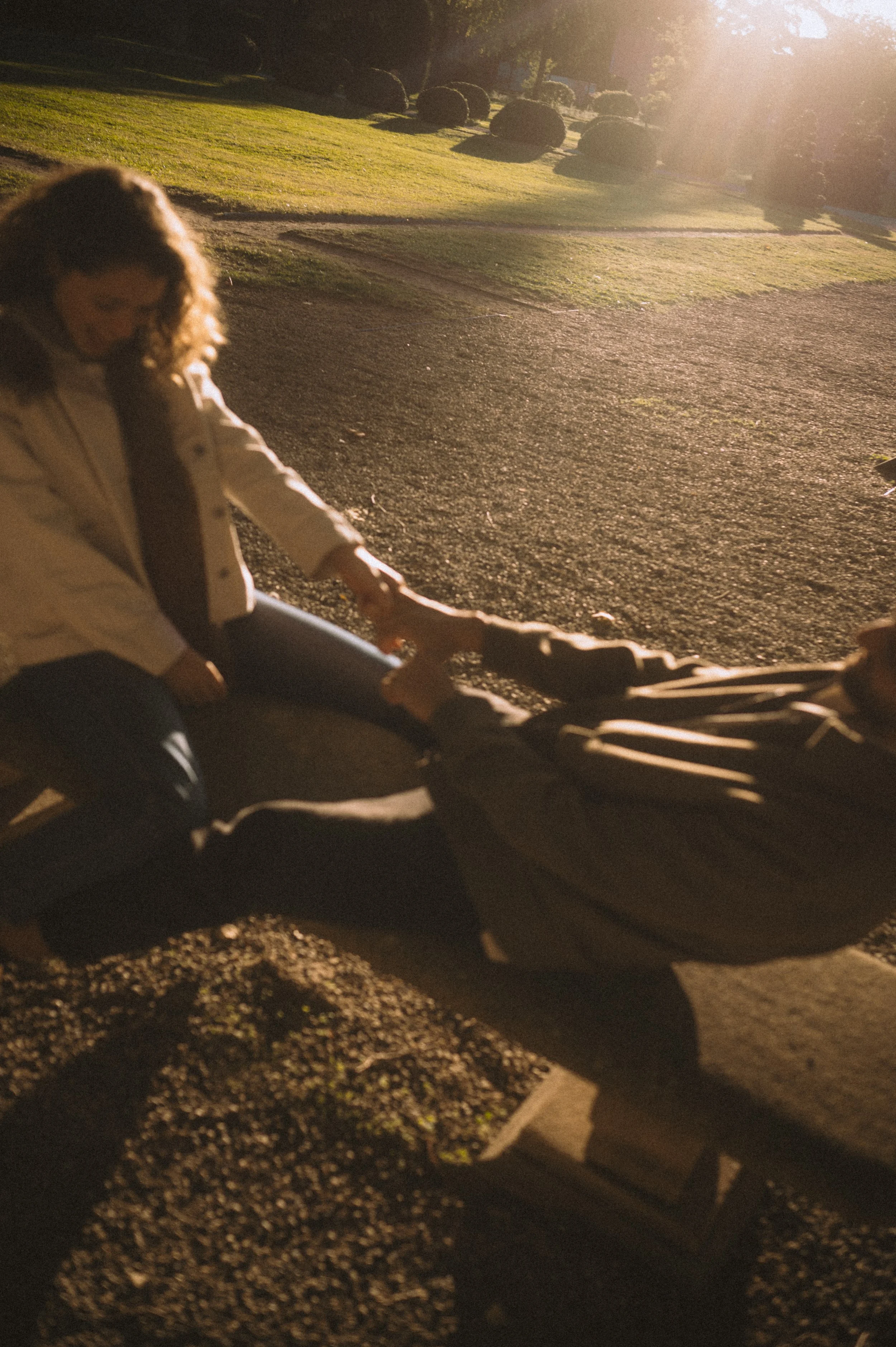 A woman with curly hair wearing a white jacket, leaning over a stroller in a park during sunset.