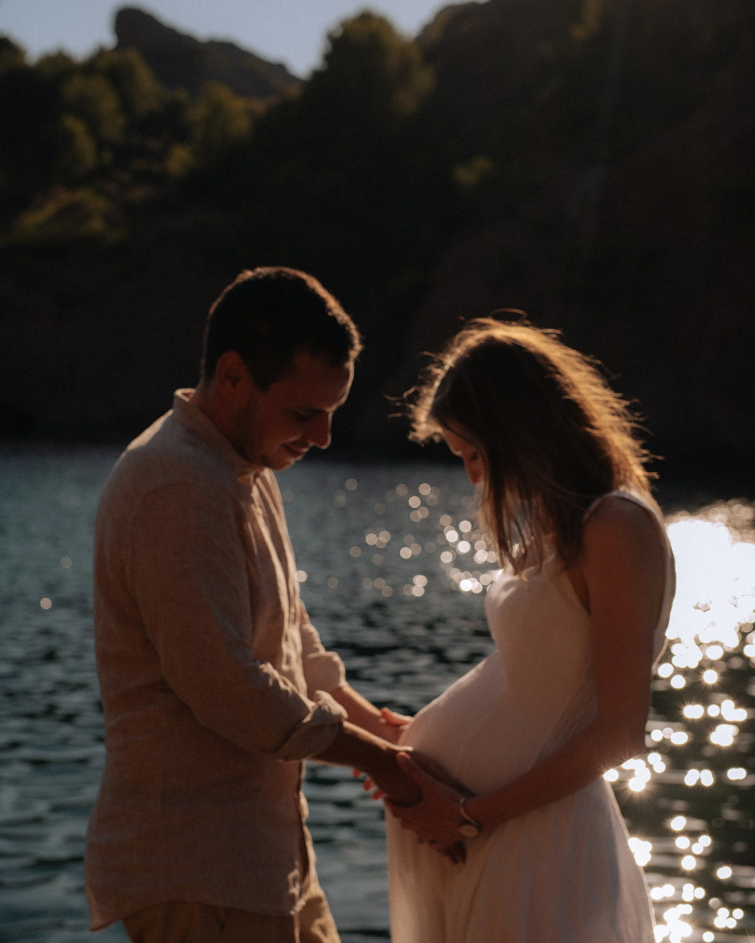 A couple standing by the water, holding hands and embracing, with the woman's pregnant belly, during sunset.