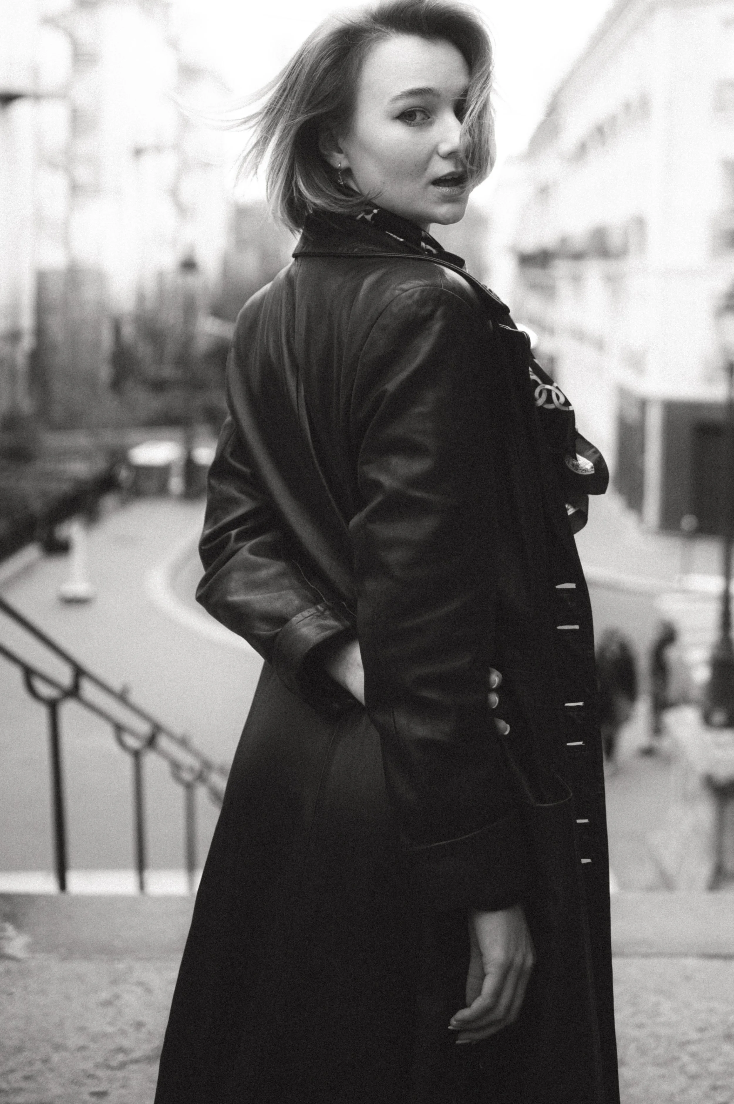 A woman with short hair posing on a city street, wearing a leather jacket, black gloves, and a patterned scarf, in black and white photography.