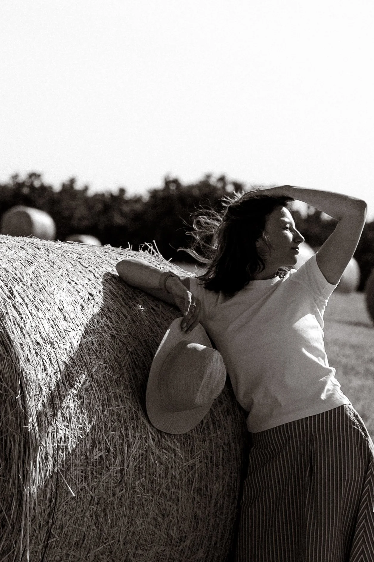 A woman leaning against a hay bale in a field, holding a hat and shielding her eyes from the sun.