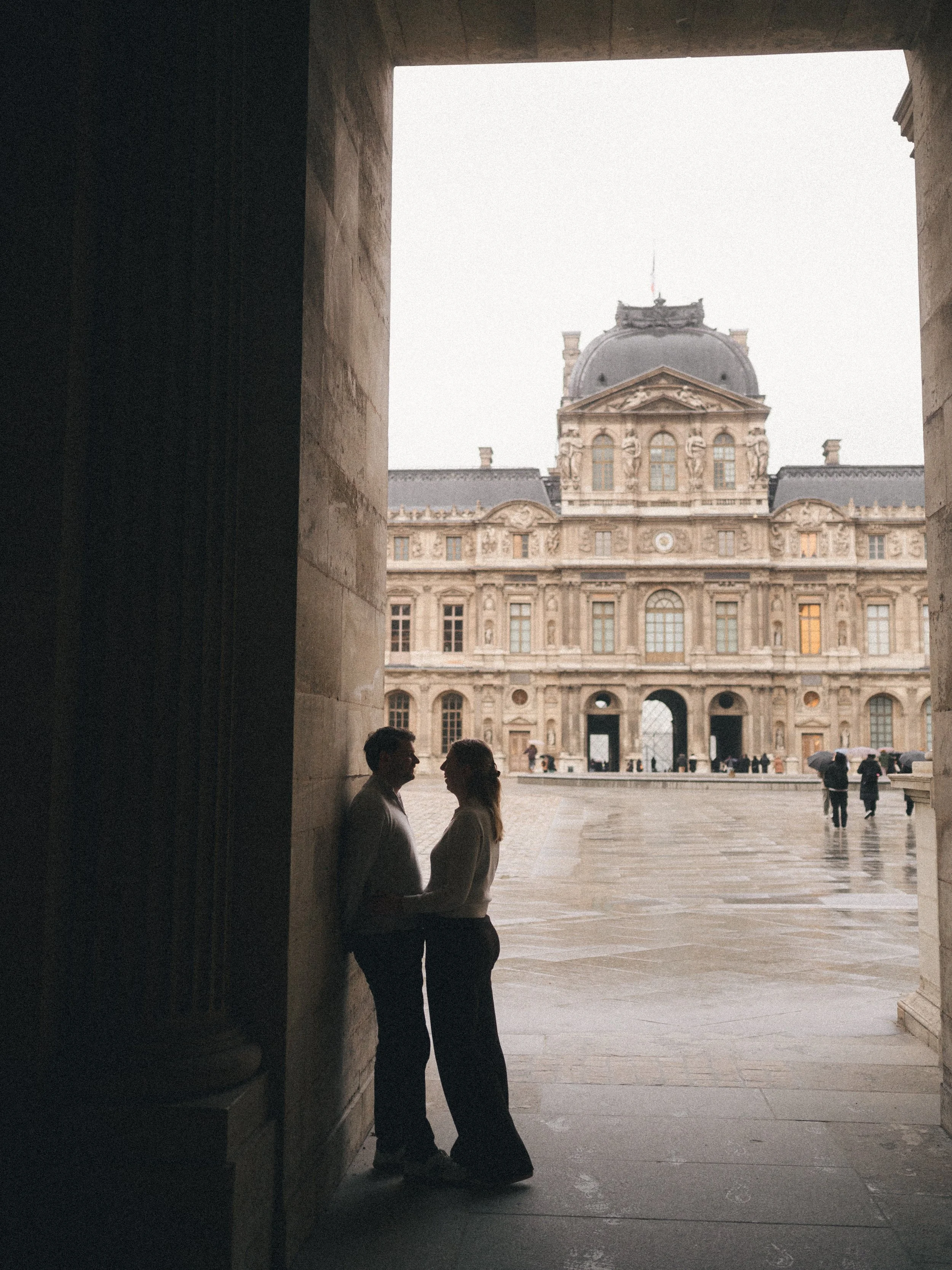 A silhouette of a man and a woman holding hands and facing each other in a stone archway with a historic building in the background on a rainy day.