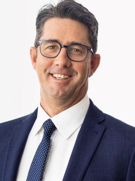 An image of Stephen Kenealy, Tech entrepreneur and AI change maker. He is a professional man with dark hair, glasses, wearing a navy suit, white shirt, and navy tie with white polka dots, smiling at the camera.