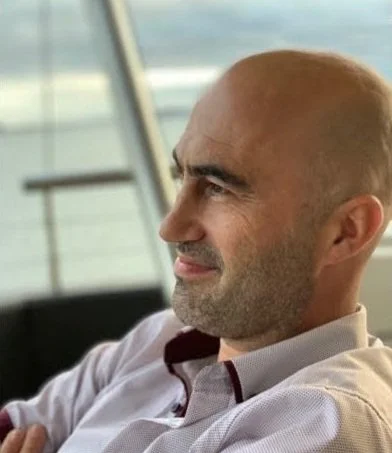 A picture of James Bisson, on of our Linkcredibles. He has a shaved head, light beard, wearing a light-colored collared shirt, sitting indoors with a blurred view of water outside a window.