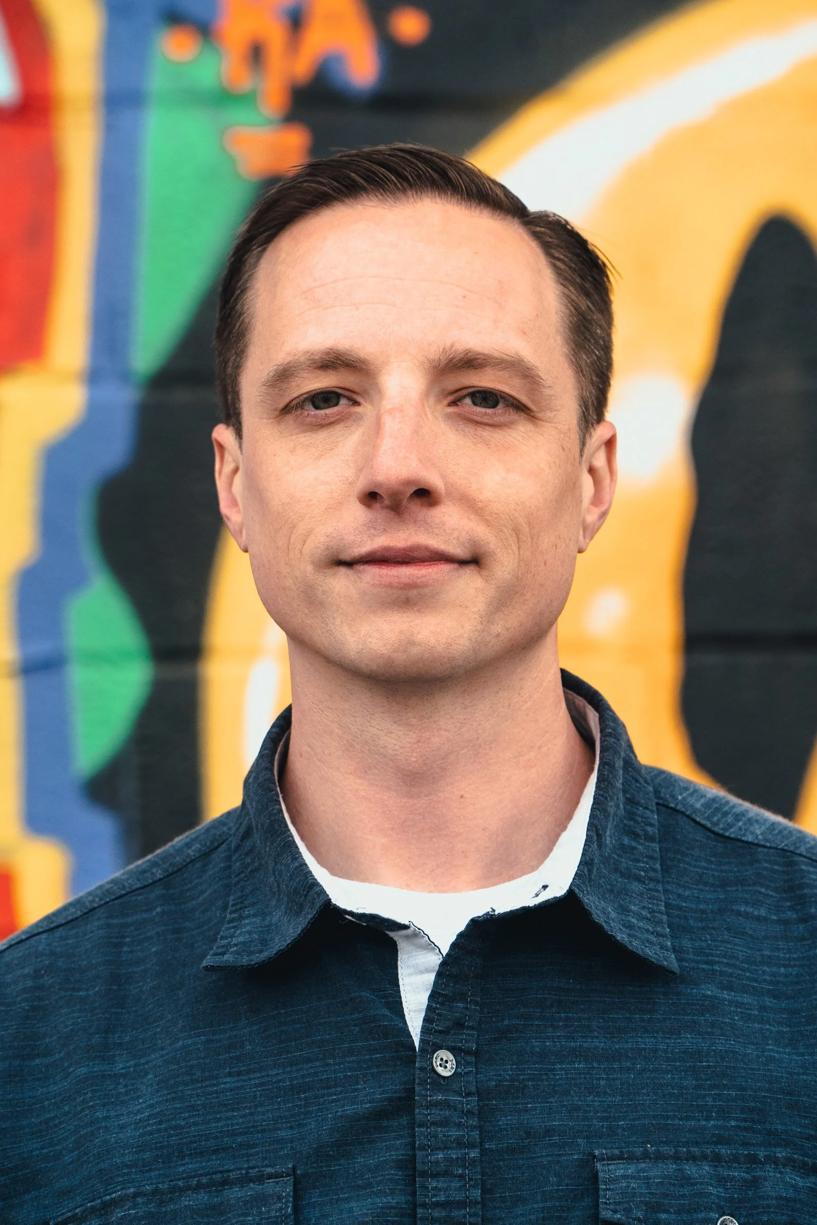 Portrait of a man with short dark hair, wearing a dark blue shirt, standing in front of a colorful graffiti wall.