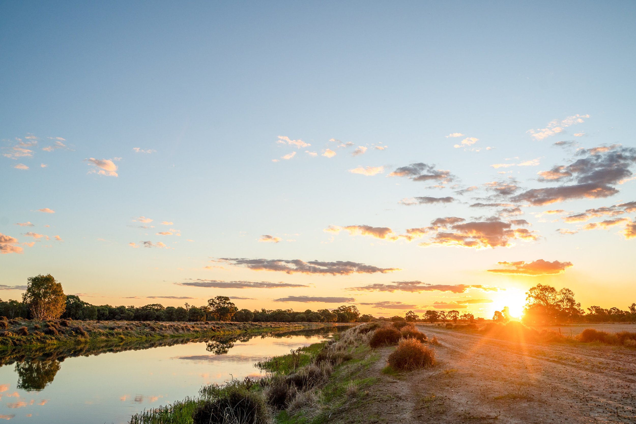 Sunset over a rural landscape with a dirt road, bushes, trees, and a calm river reflecting the sky, with clouds and orange sunlight