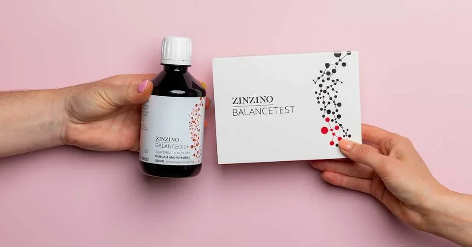 A person holding a bottle labeled "Zinzino Balance" and another person holding a white box with the same branding and a black and red botanical design, against a pink background.