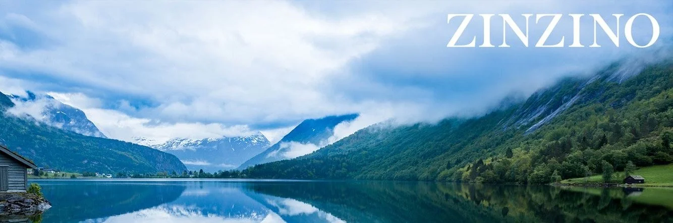 Serene mountain lake landscape with snow-capped peaks, lush green forested hills, a small house by the water, and cloudy skies. The word "ZINZINO" is written in white text at the top right corner.