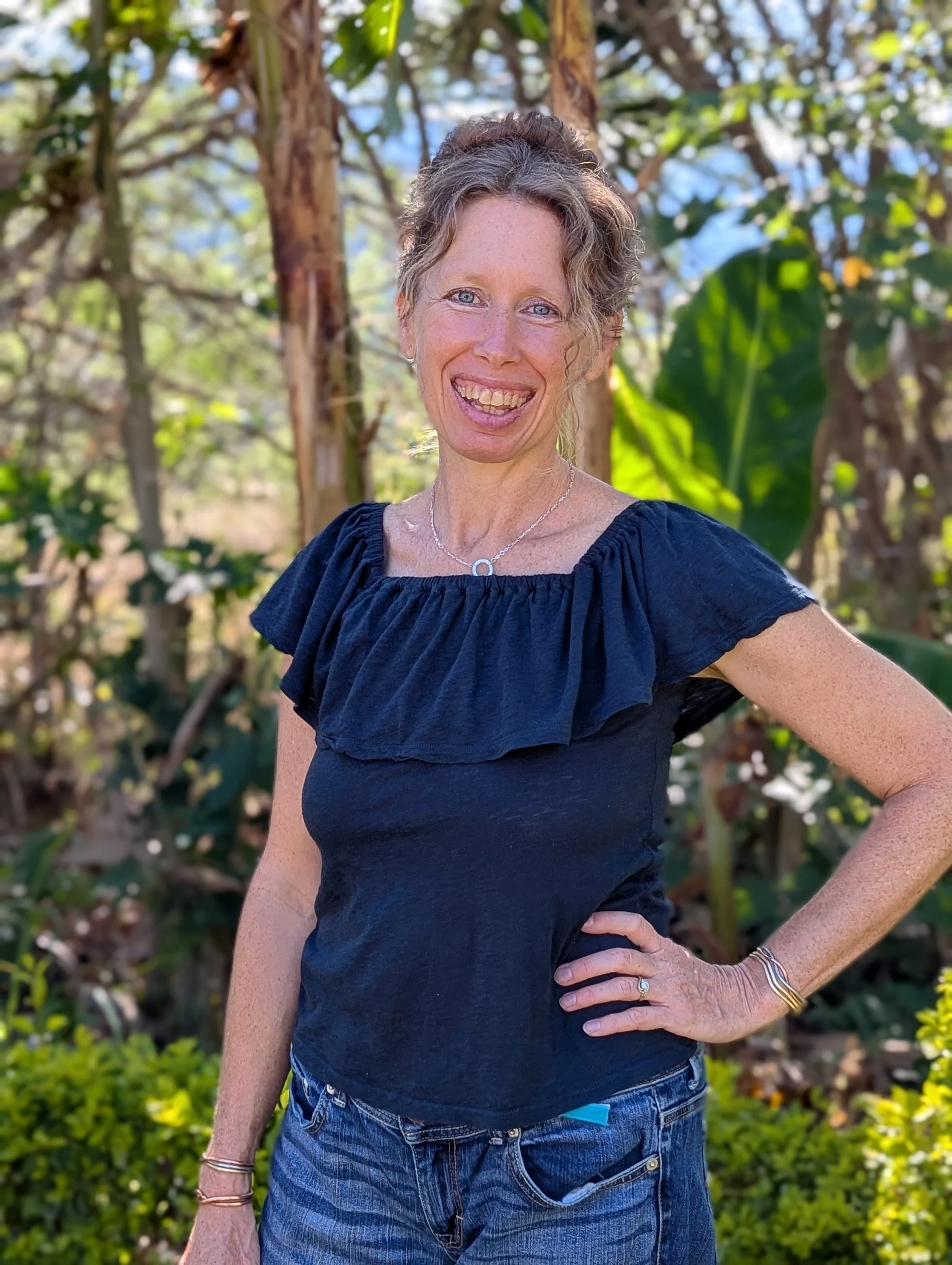 A woman with curly hair and freckles smiling outdoors in a garden or park, wearing a black top, jeans, and jewelry, with trees and green foliage in the background.