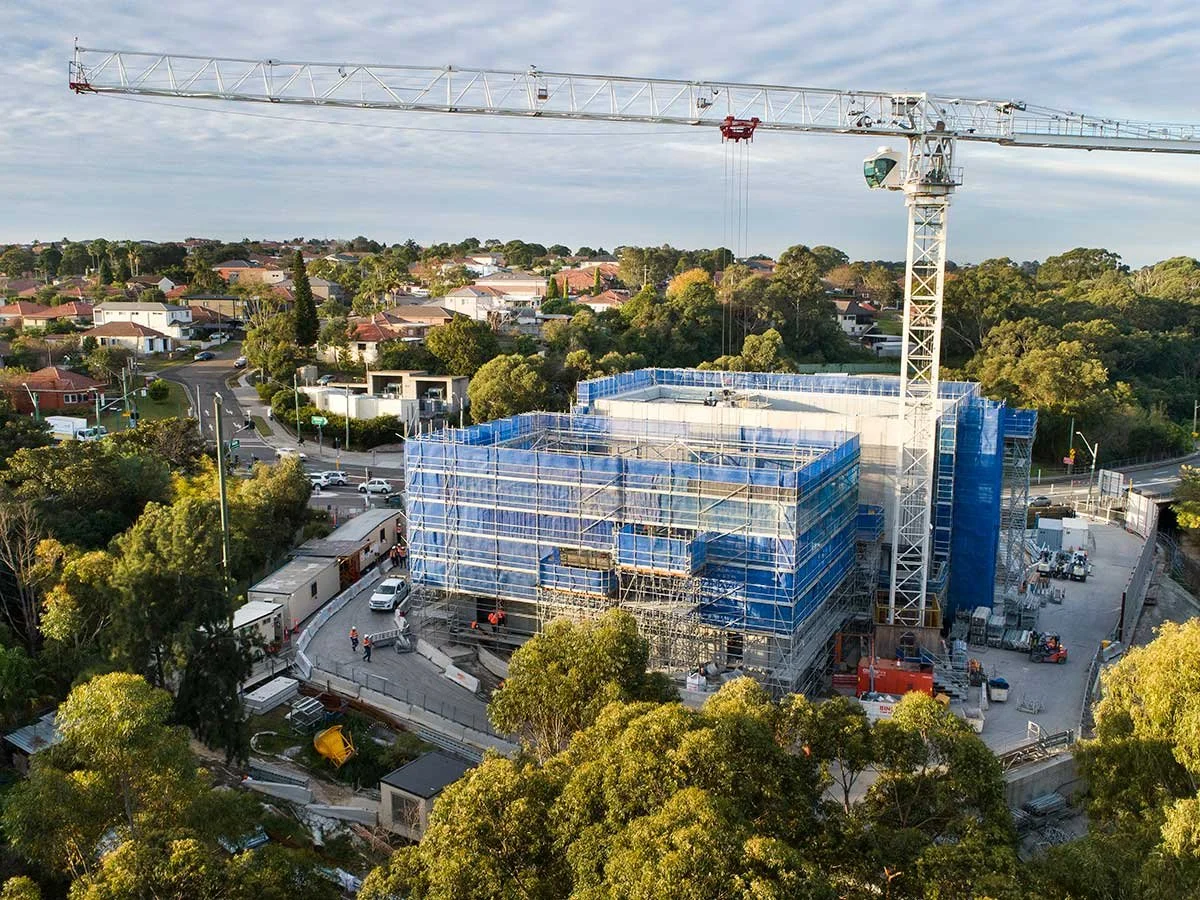 Construction site with a building covered in blue scaffolding, a tall crane, and surrounded by trees and residential houses.