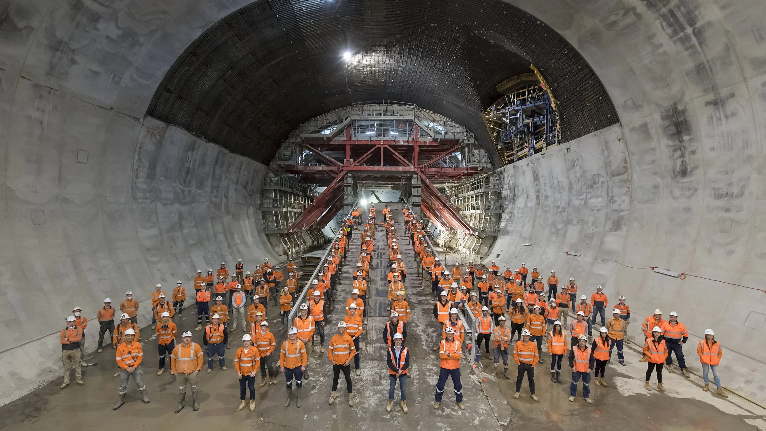Group of construction workers in orange safety jackets and white helmets inside a large tunnel under construction, posing for a photo.