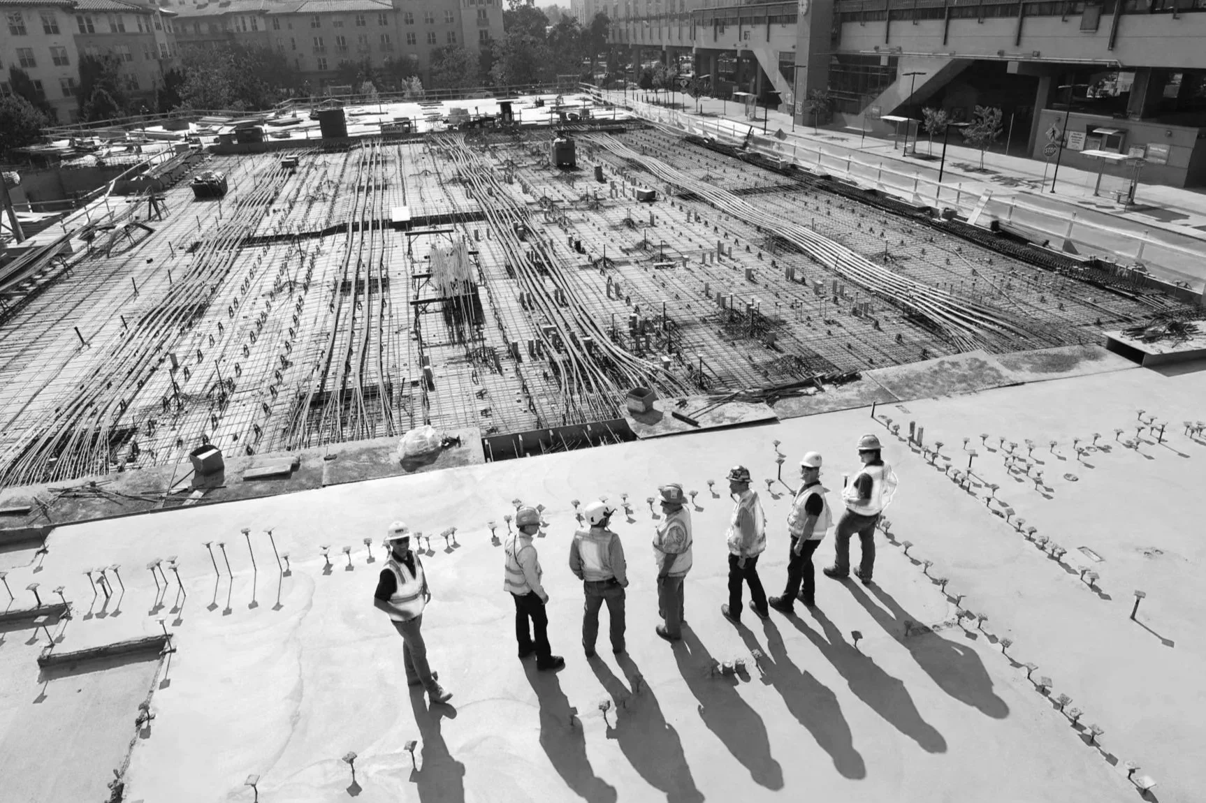 Group of construction workers wearing safety vests and helmets standing on concrete rooftop observing an extensive building construction site with numerous steel reinforcements and utility lines.