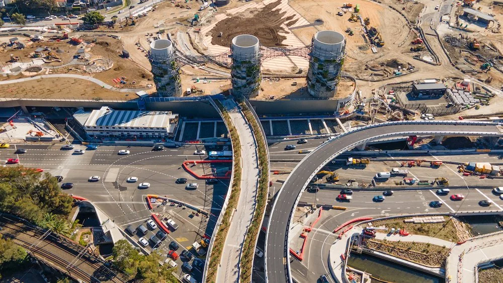 An aerial view of a multi-level highway interchange with cars, construction sites, and industrial structures under development.