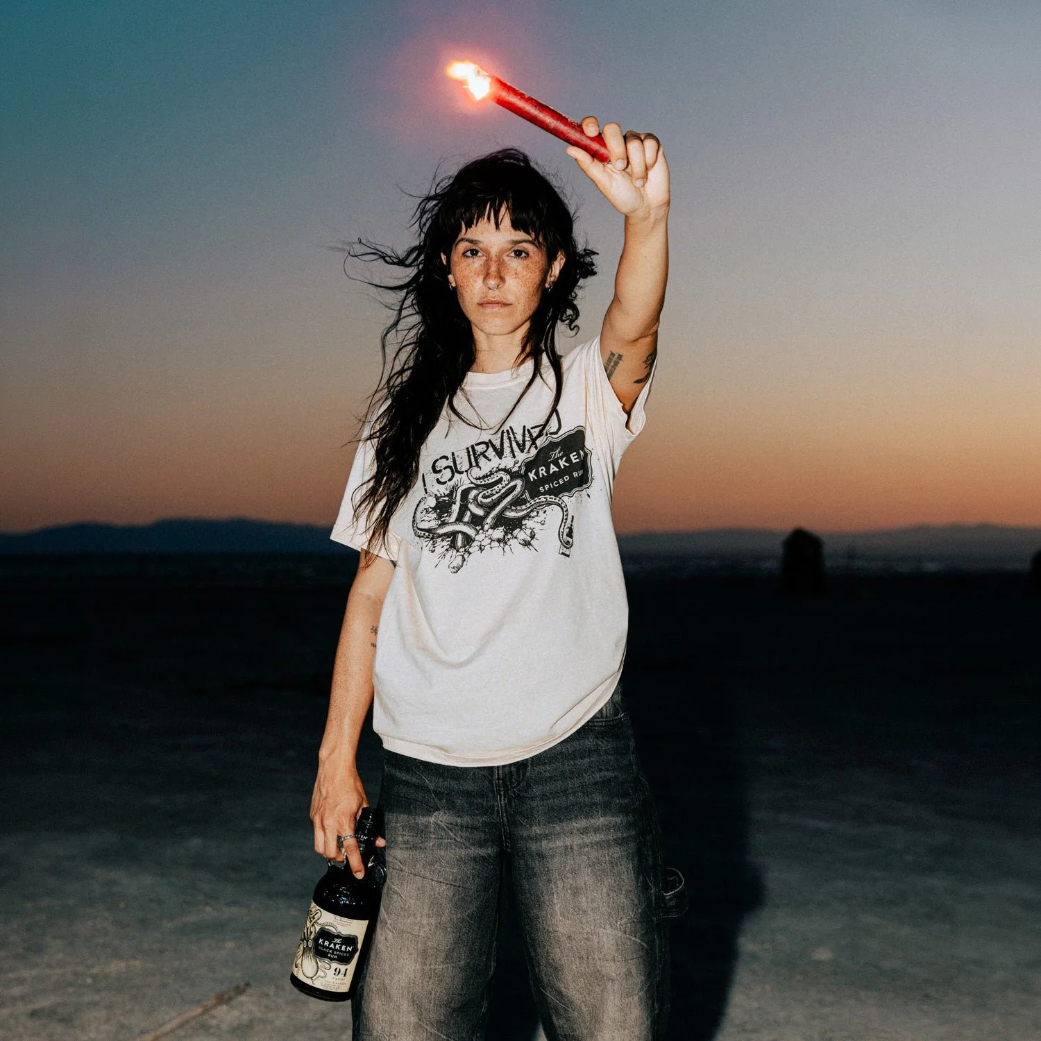 A young woman with dark hair and freckles is holding a bottle of Kraken rum in her left hand and a lit firecracker in her right hand. She is wearing a white T-shirt with a graphic design and text that says 'Survivor the Kraken Spiced Rum.' The background shows a sunset or dusk at a beach or desert, with a dark landscape and a few rocks or formations in the distance.