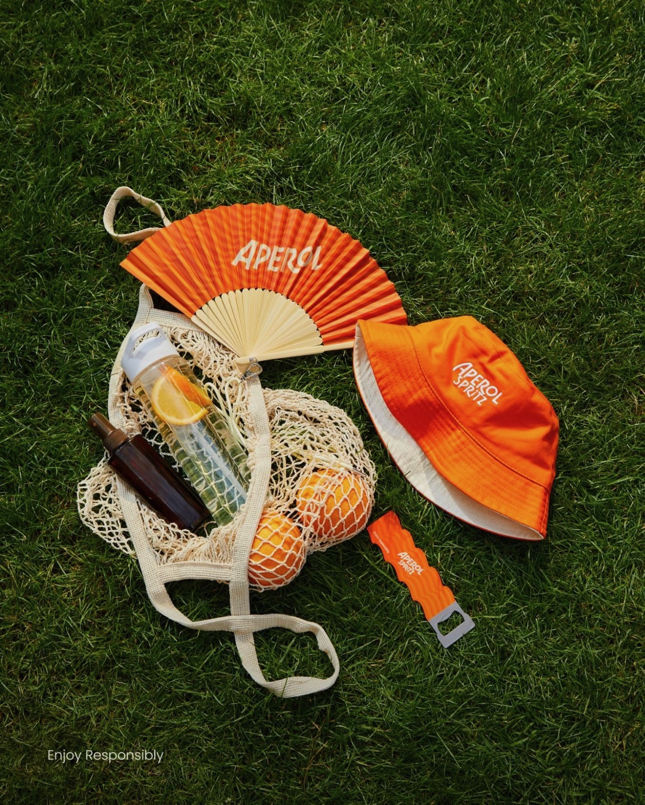 A collection of orange-themed summer items on grass includes a hand fan, a hat, a water bottle, and a mesh bag with oranges.