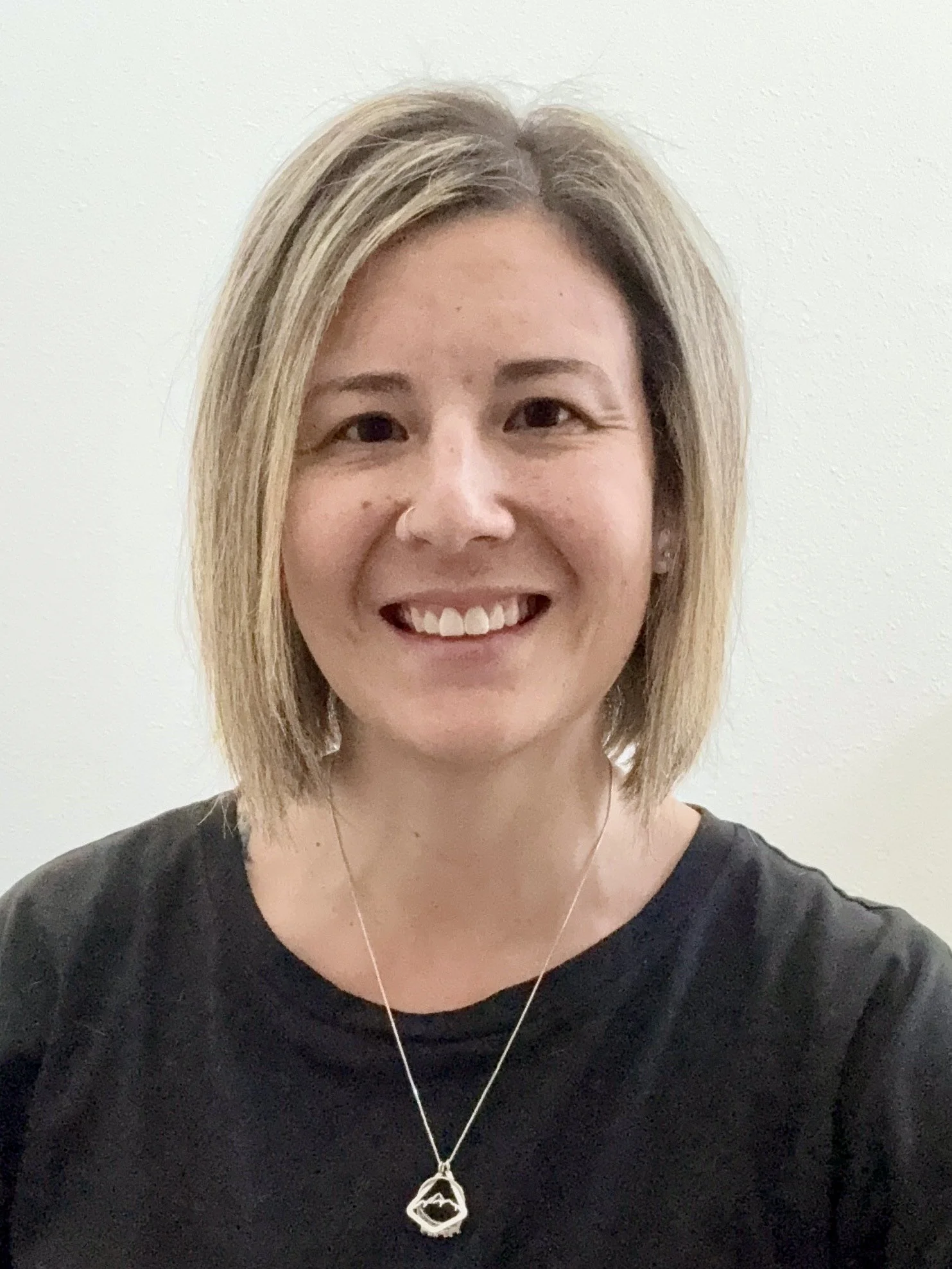 A woman with shoulder-length blonde hair smiling at the camera, wearing a black shirt and a necklace with a geometric pendant against a plain light-colored wall.
