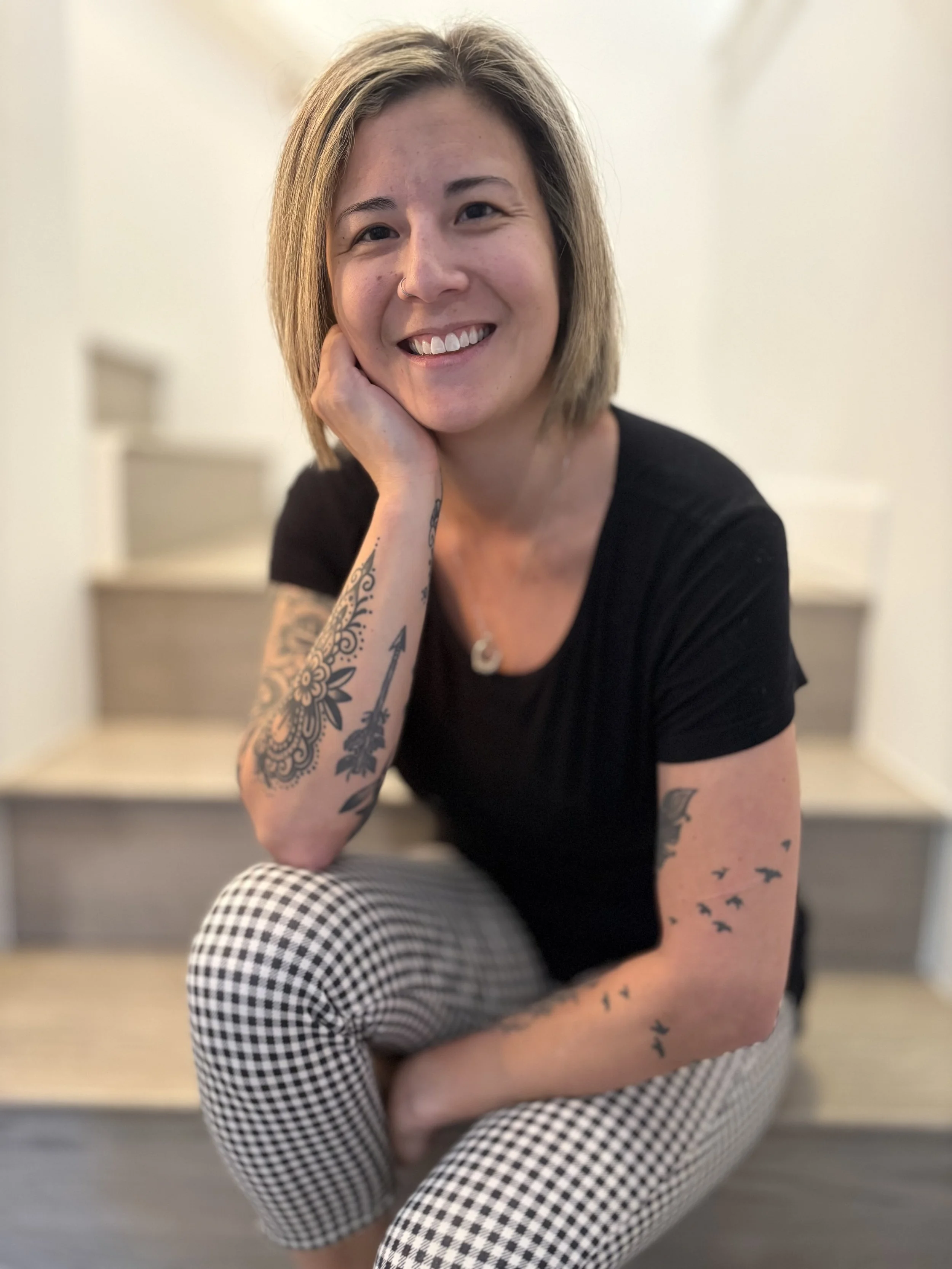 A smiling woman with short hair, tattoos on her arm, wearing a black shirt and checkered pants, sitting on steps.