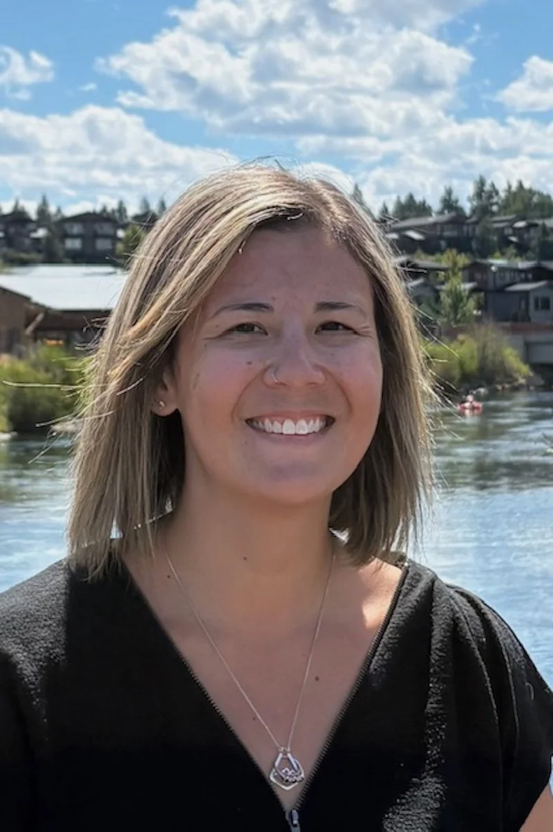 A female trauma therapist in Bend, OR with shoulder-length blonde hair standing outdoors near a body of water, with houses and trees in the background under a cloudy sky.