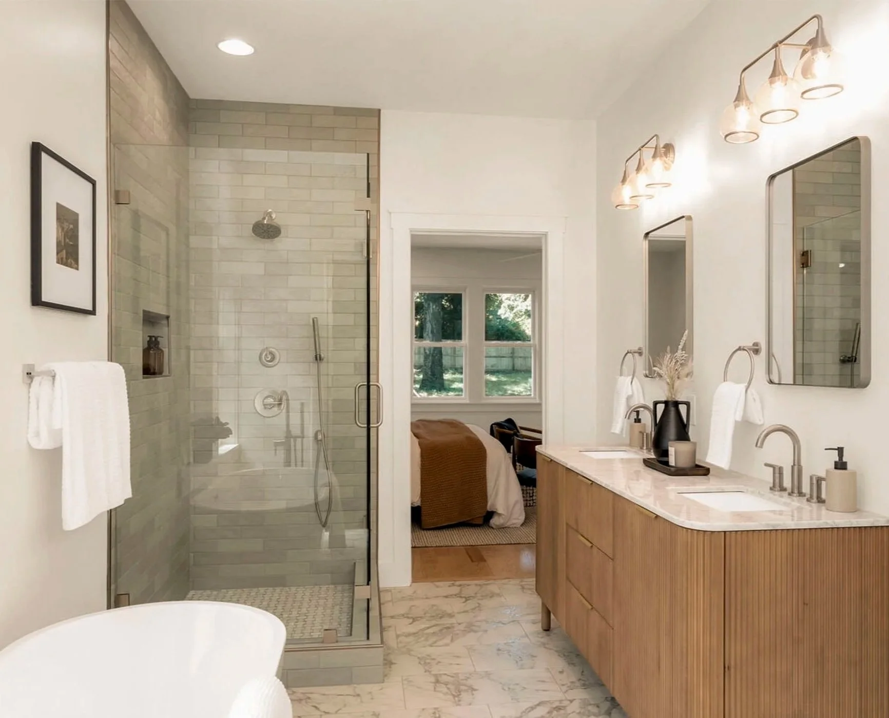 Modern bathroom with a glass shower enclosure, double vanity with two sinks, mirrors, and light fixtures. There is a window showing a yard outside, and a bed can be seen through the open door.
