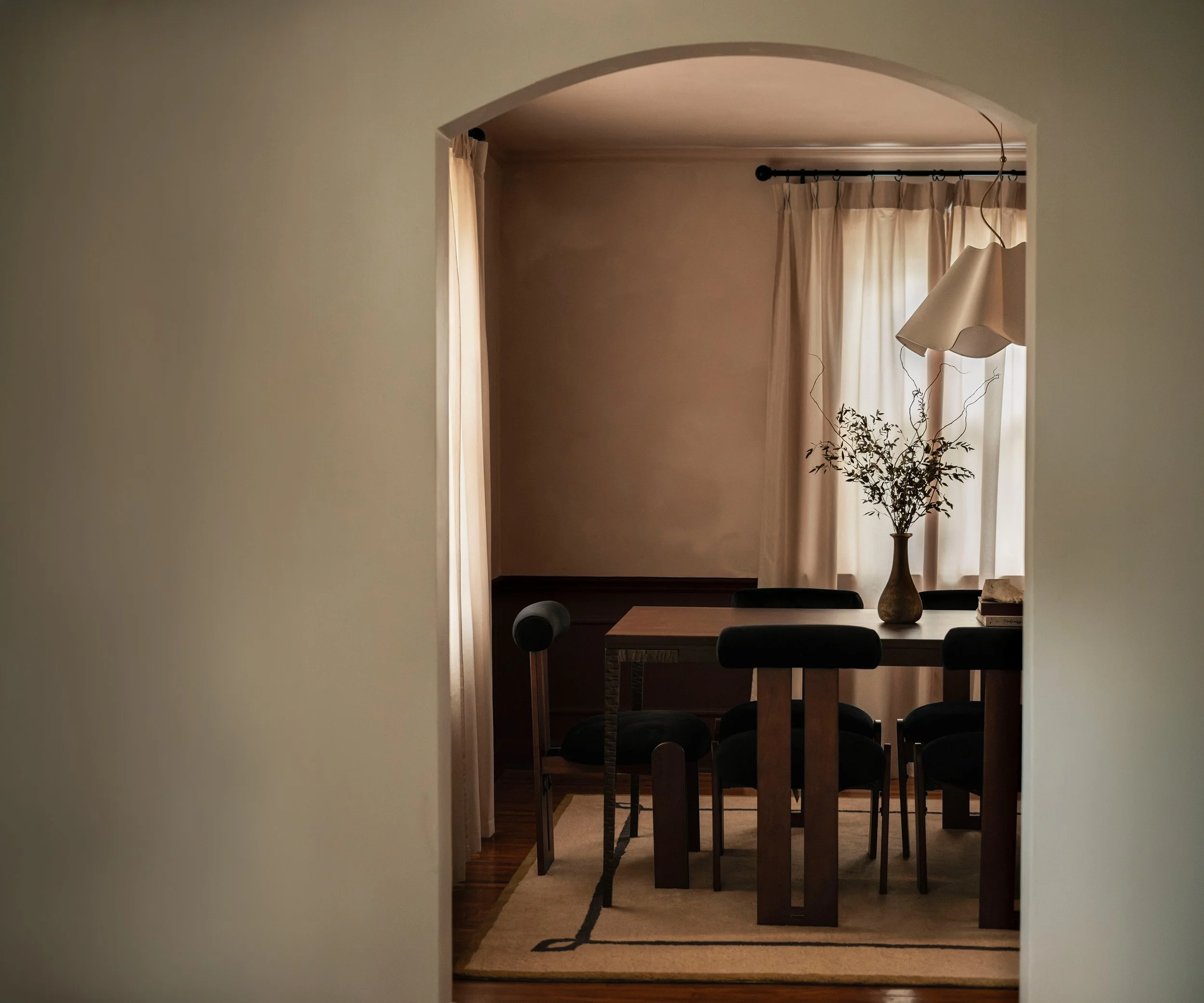 Dining room with a rectangular wooden table, six black chairs, a decorative vase with branches, curtains, and a hanging light fixture.