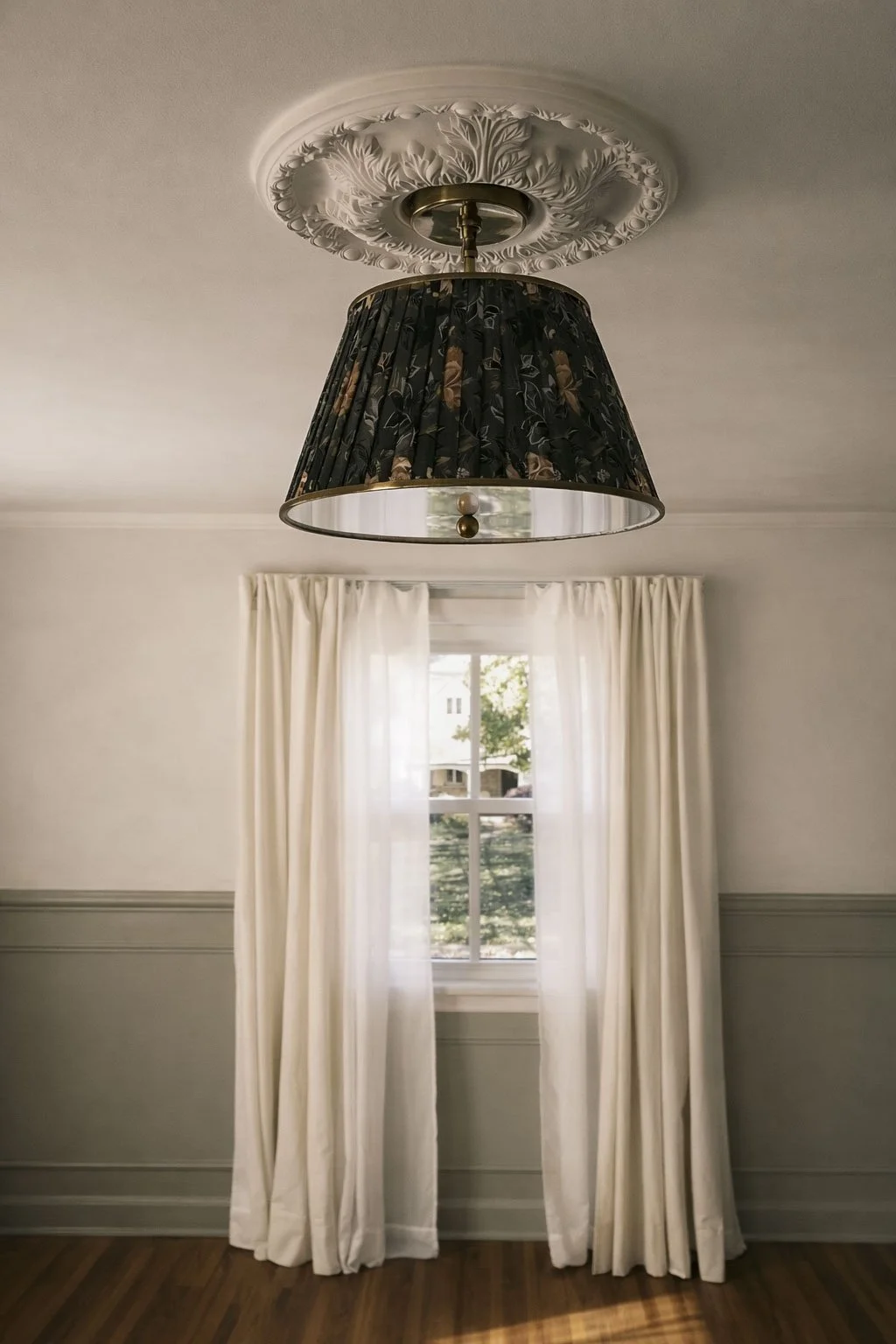 A ceiling light fixture with a decorative floral ceiling medallion, a black floral lampshade hanging from the ceiling, and a window with white curtains allowing natural light into a room with white walls and hardwood floors.
