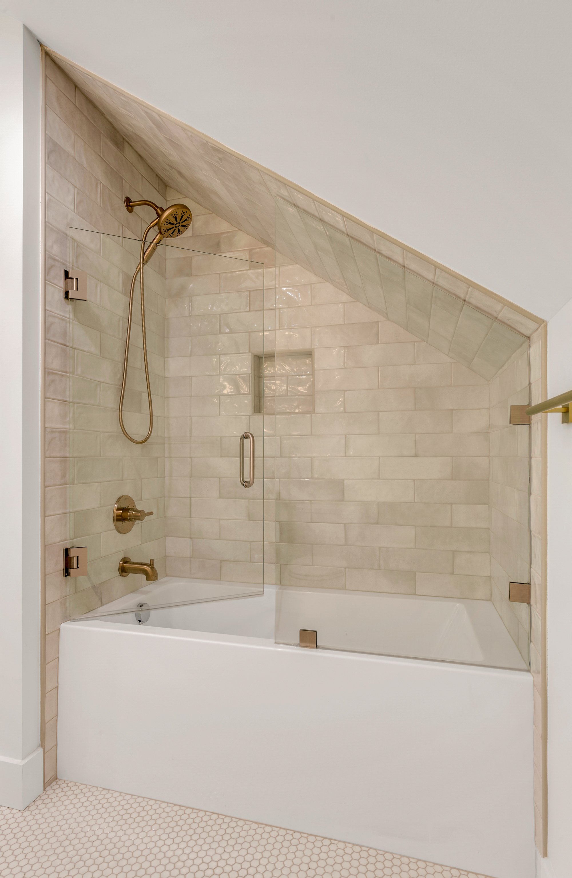 Bathroom with a walk-in shower featuring beige subway tile walls, a glass door, gold fixtures including a showerhead and handle, and a white bathtub. The floor has small hexagon tiles.