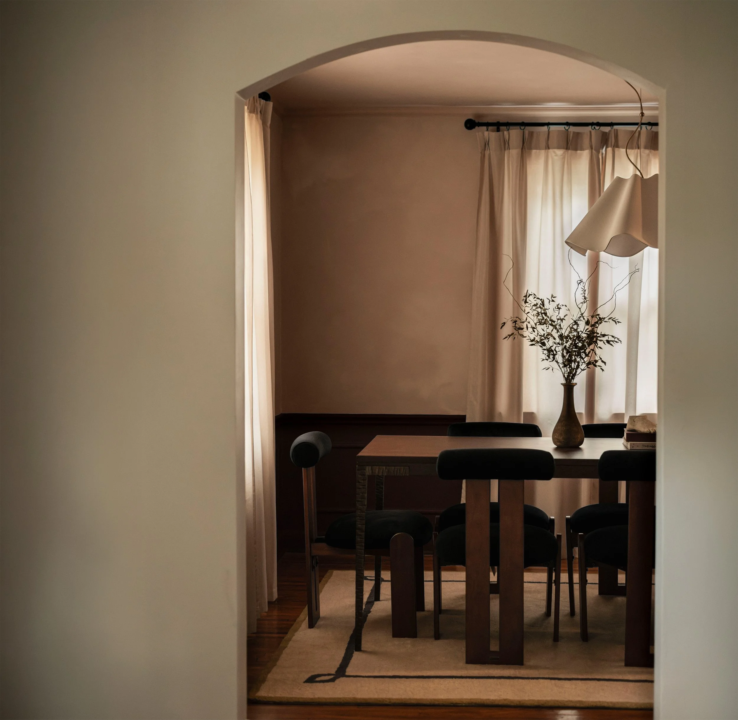 A view into a dining room through an archway, with a wooden table, black chairs, a beige carpet with black trim, a large beige curtain, and a tall vase with branches and dried plants.
