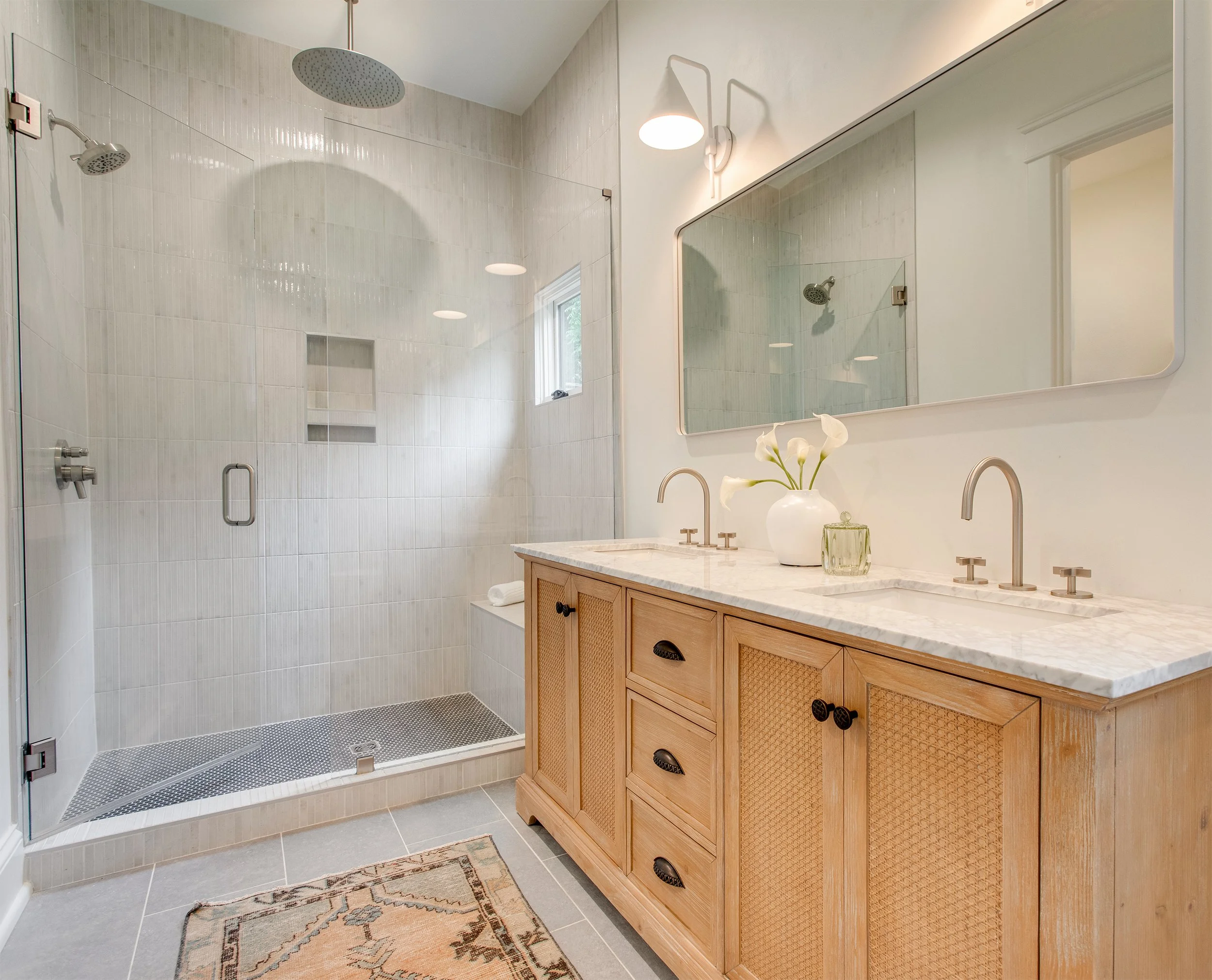 Bathroom with a walk-in shower enclosed by glass, with a rainfall showerhead and hand-held shower, a wooden vanity with a marble countertop, a mirror, and decorative vase with white flowers