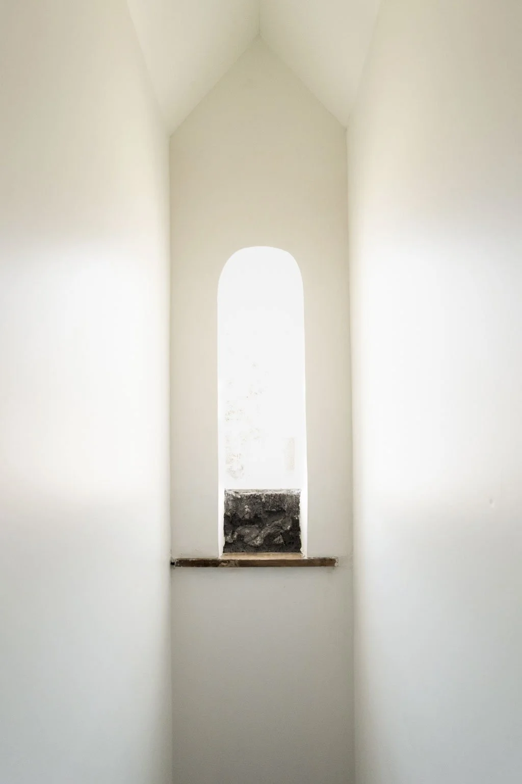 Interior view of a narrow arched window with white walls and a small opening at the bottom showing rocks and dirt outside.