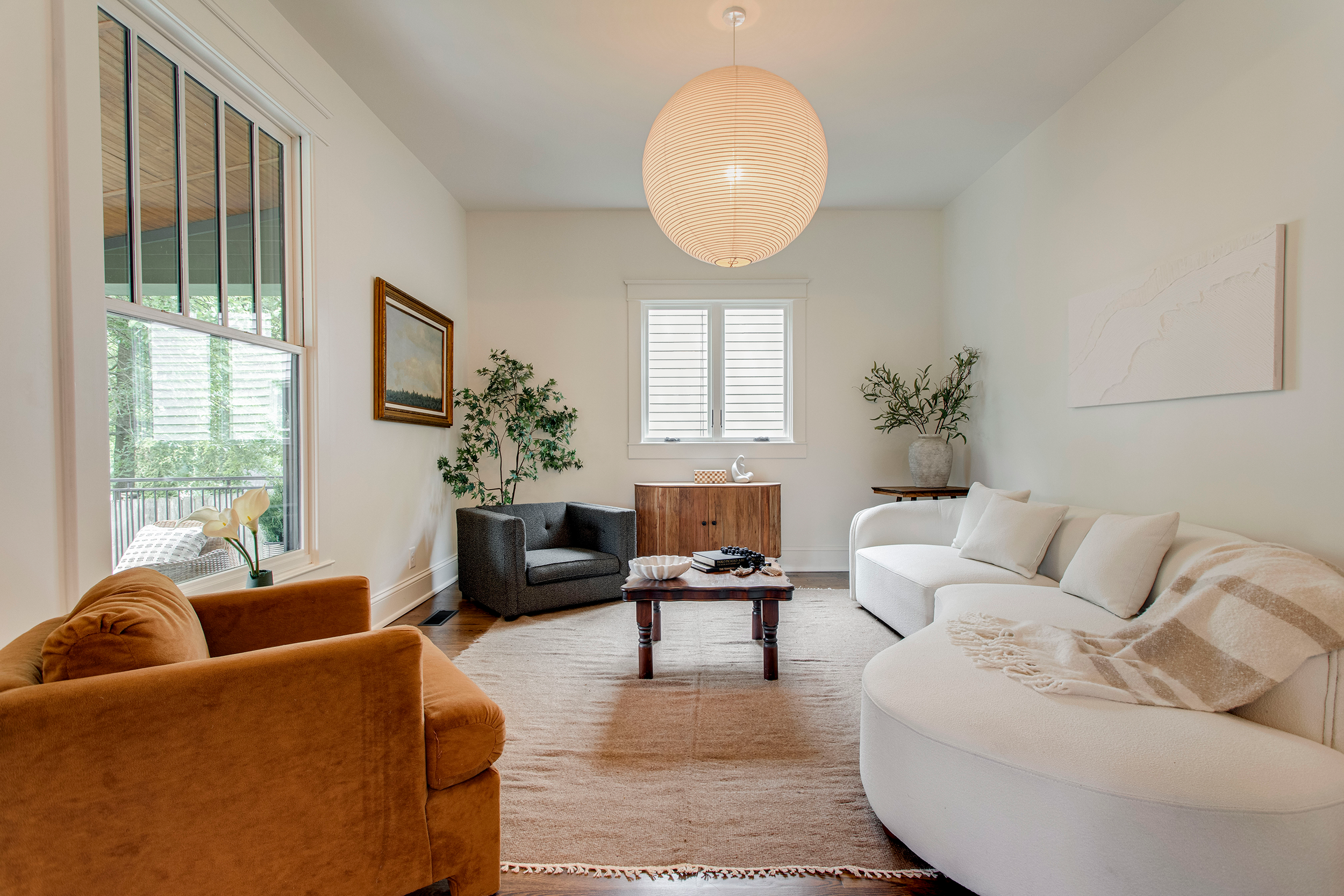 Living room with beige walls, large windows, and a white ceiling. Features include a rust-colored armchair, a gray armchair, a white curved sofa, a wooden coffee table, potted plants, wall art, and a large paper lantern overhead.