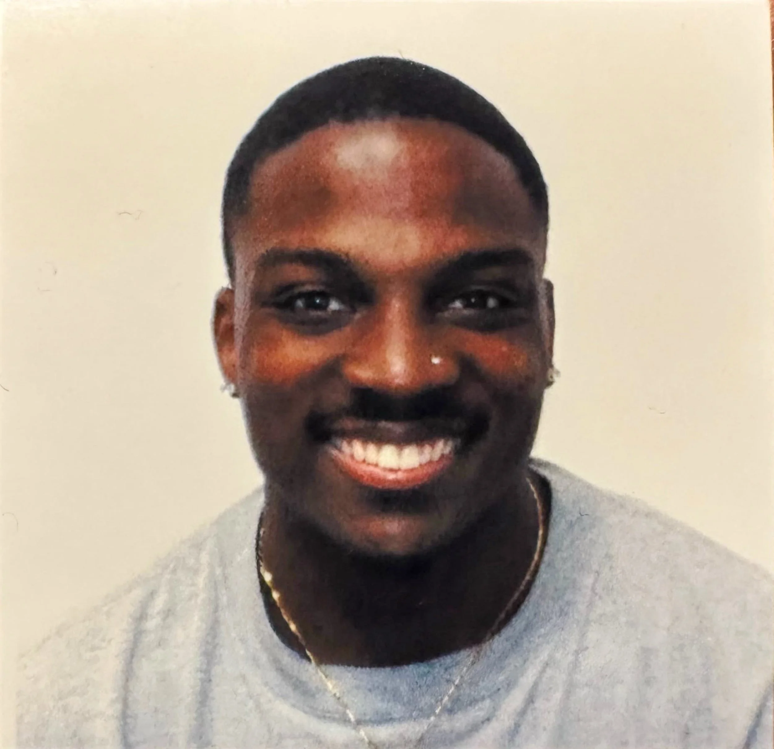 Close-up photo of a young man with short hair, wearing a gray shirt, smiling, showing his teeth, and wearing earrings, a nose ring, and a chain necklace, against a plain light-colored background.