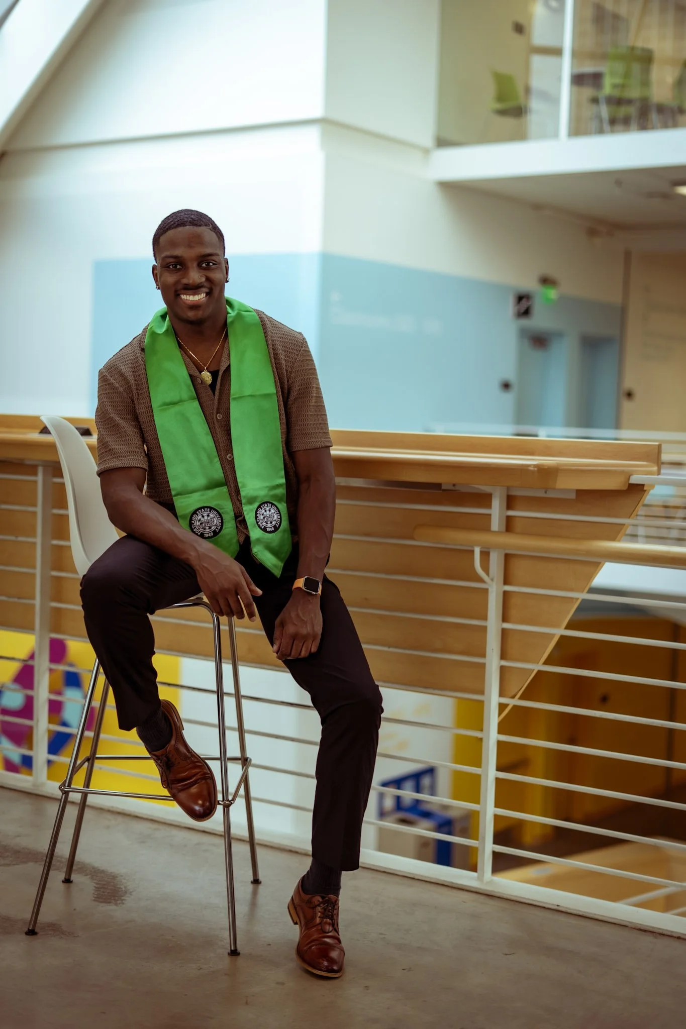 A young man in a brown shirt and black pants with a green graduation stole, smiling and sitting on a high stool in a modern building with a wooden railing and colorful decor.