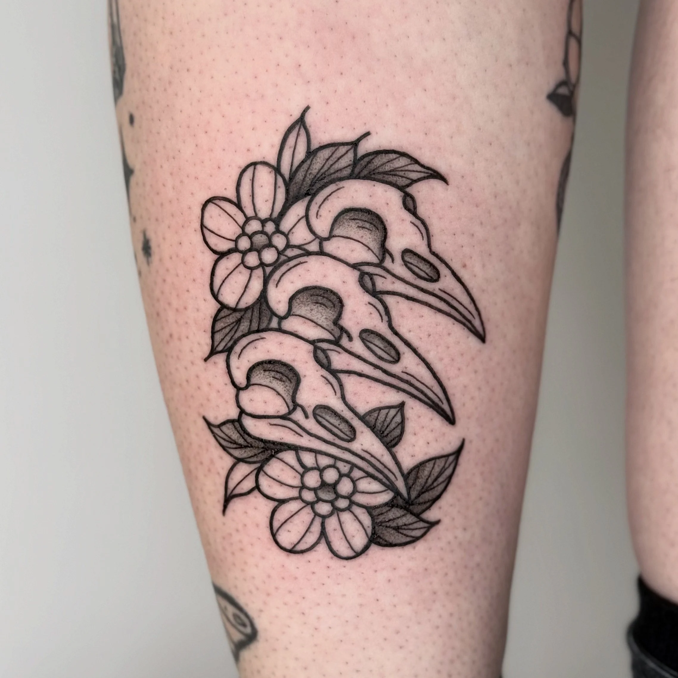 Tatouage noir et blanc représentant des fleurs et des feuilles avec trois crânes d'oiseaux stylisés.