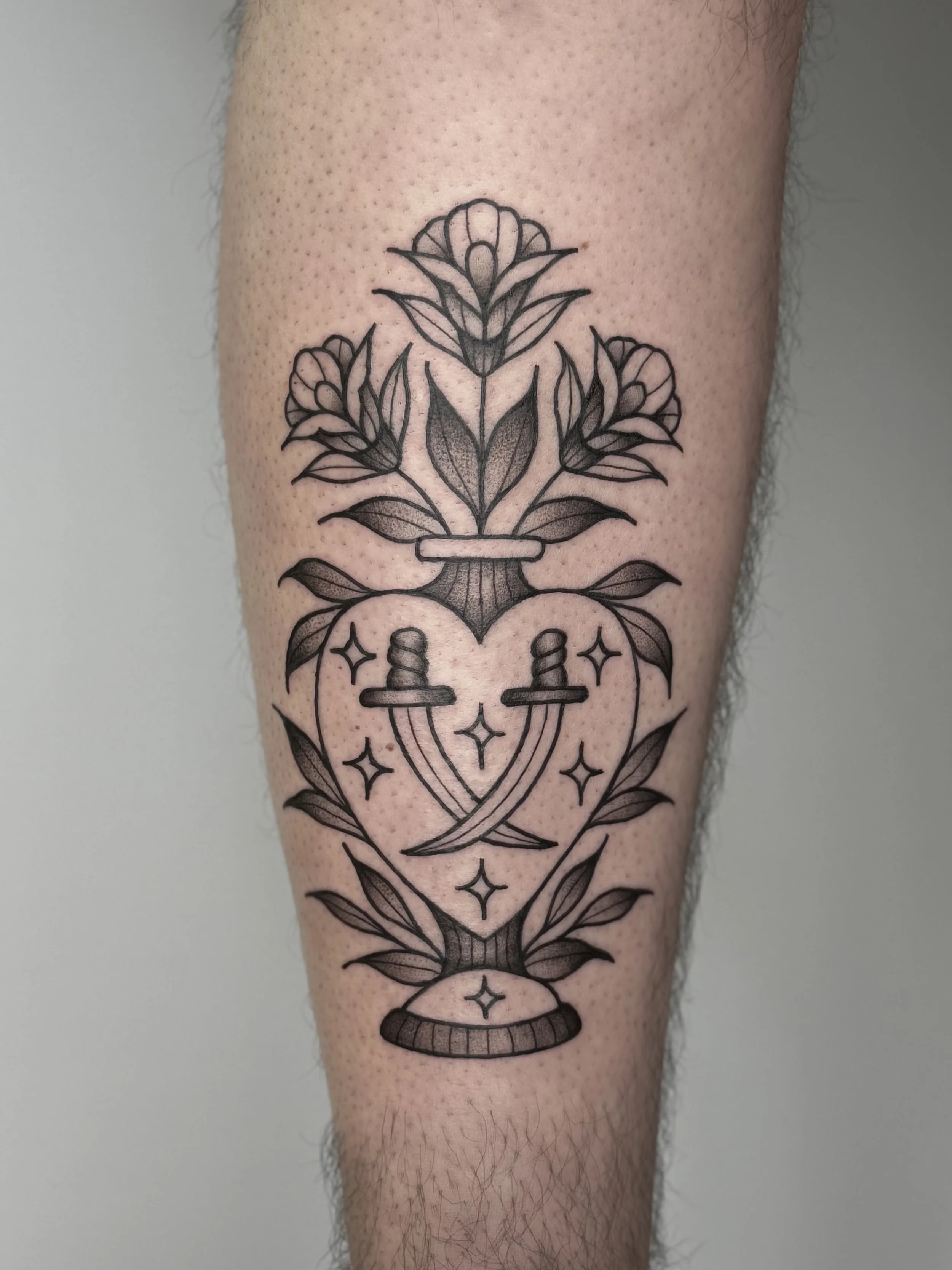 Tatouage noir et gris d'une plante avec trois fleurs, deux épées croisées dans un cœur entre les fleurs, entouré de feuilles, avec des étoiles dessinées autour.
