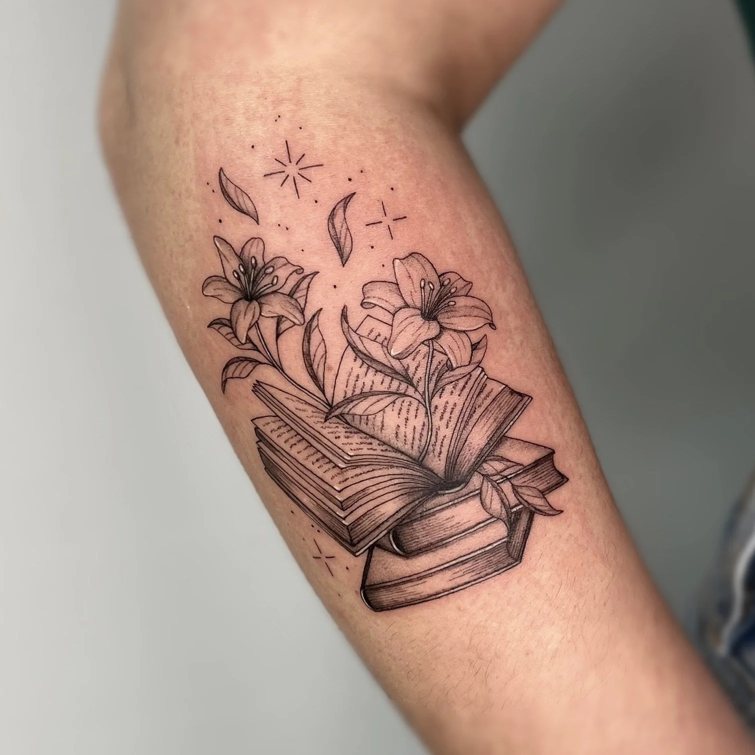 Tatouage de livres ouverts avec des fleurs lilies et des feuilles, entouré d'étoiles et de petits points, en noir et gris.