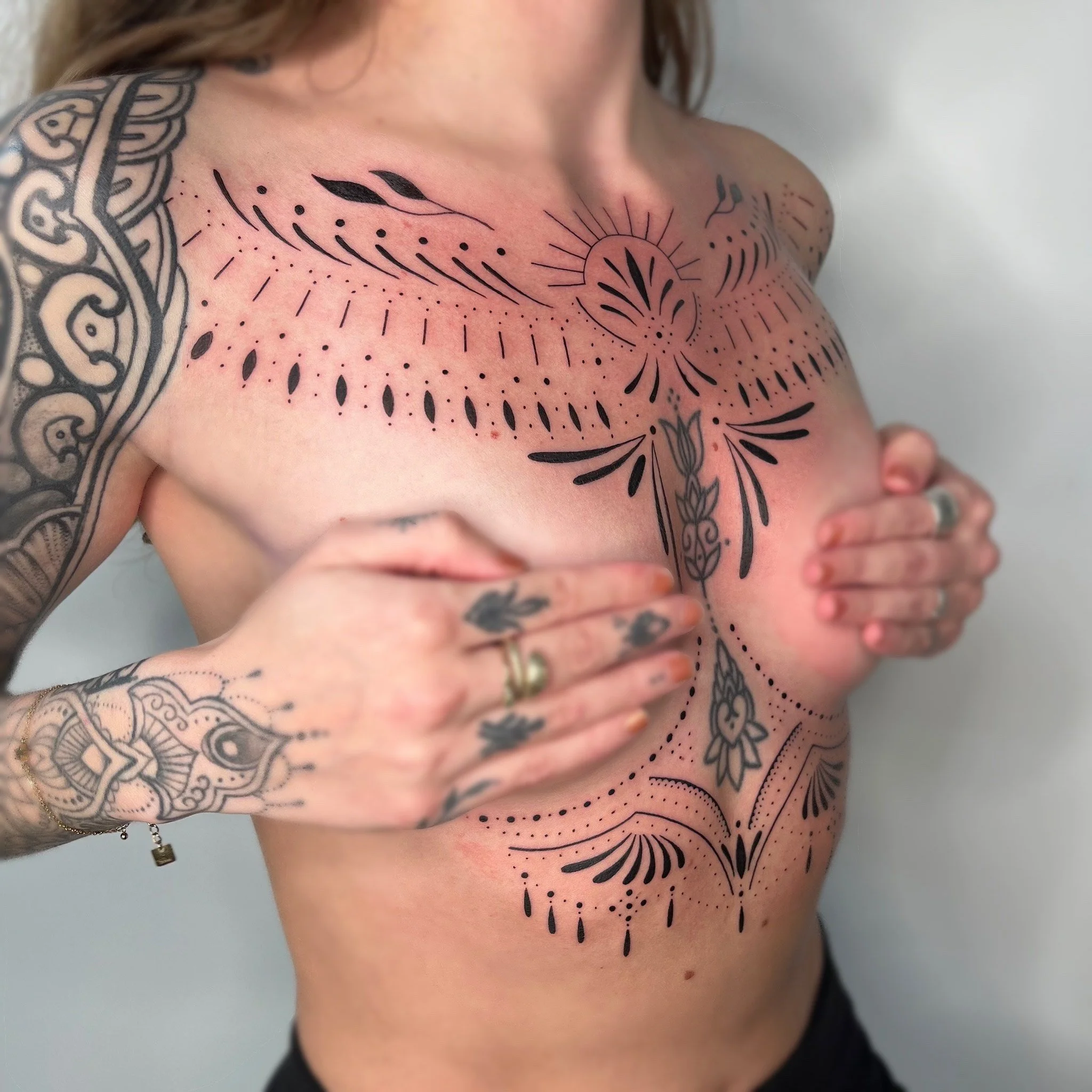 Une femme avec des tatouages noirs couvrant le haut du corps, tenant ses seins avec ses mains, et un fond blanc.