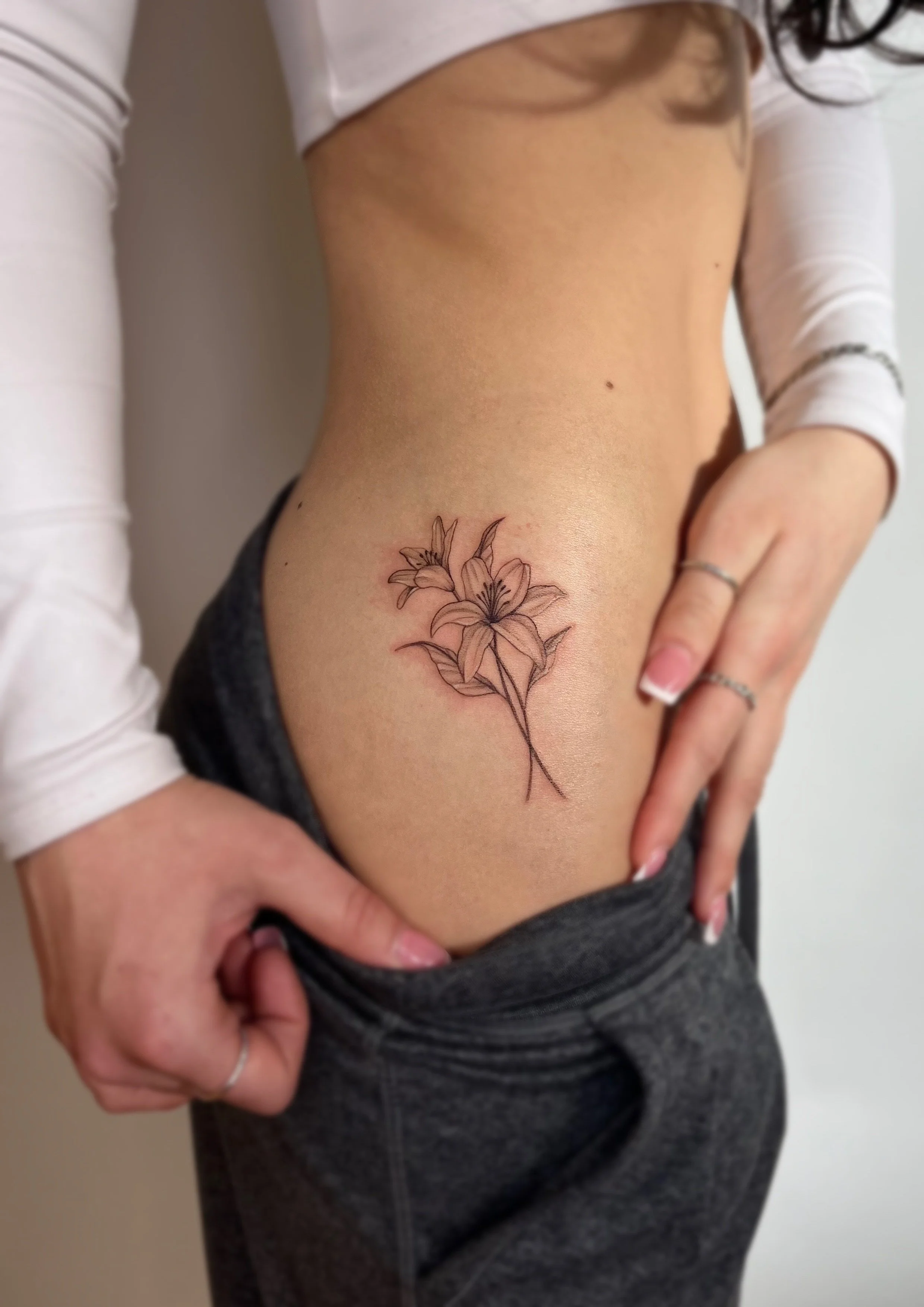 Tatouage d'une fleur d'ornithogale sur la côte d'une femme, visible à côté de ses hanches, avec un pantalon gris et un haut blanc.