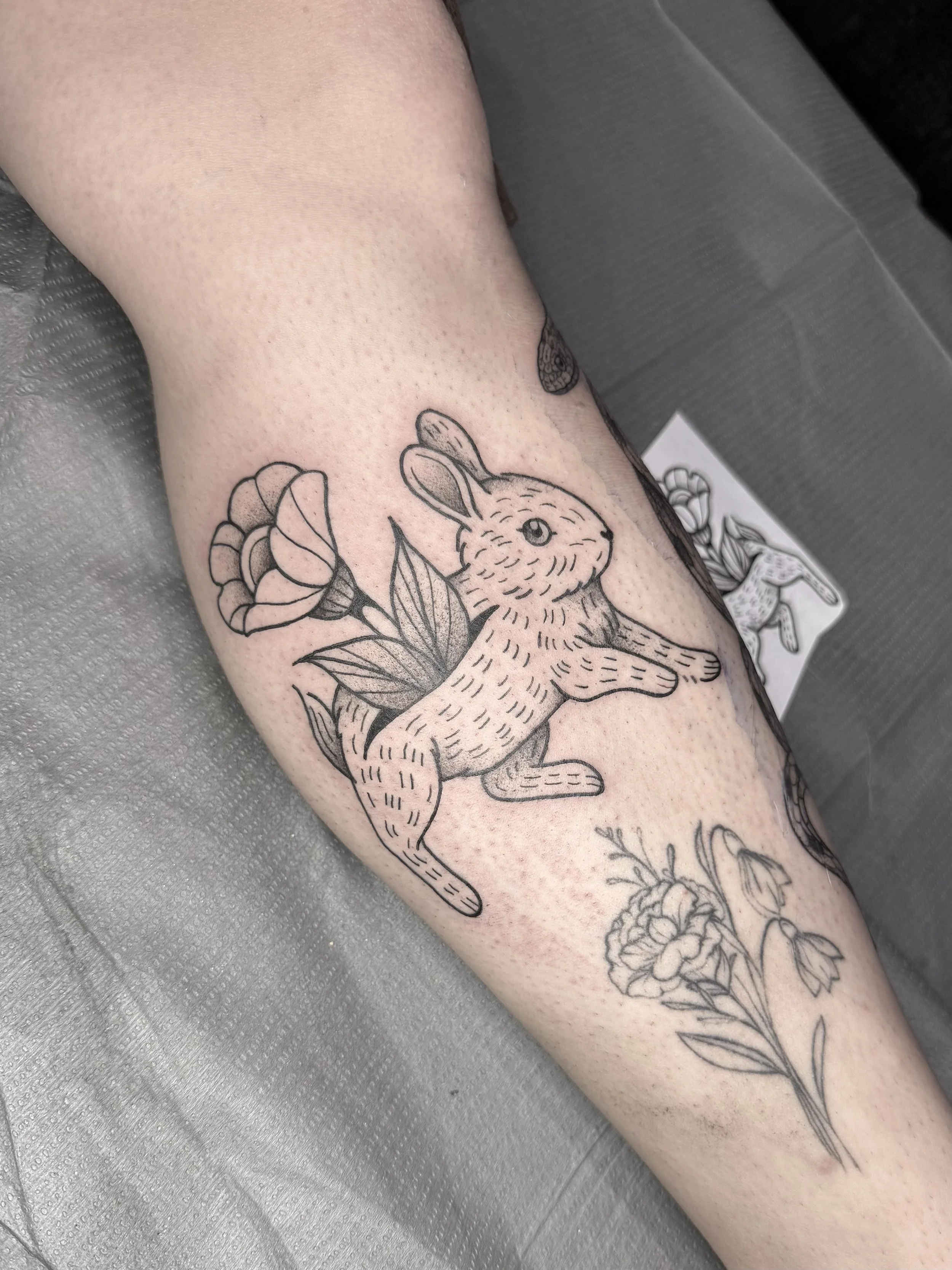 Tatouage en noir et blanc d'un lapin entouré de fleurs sur la jambe d'une personne.